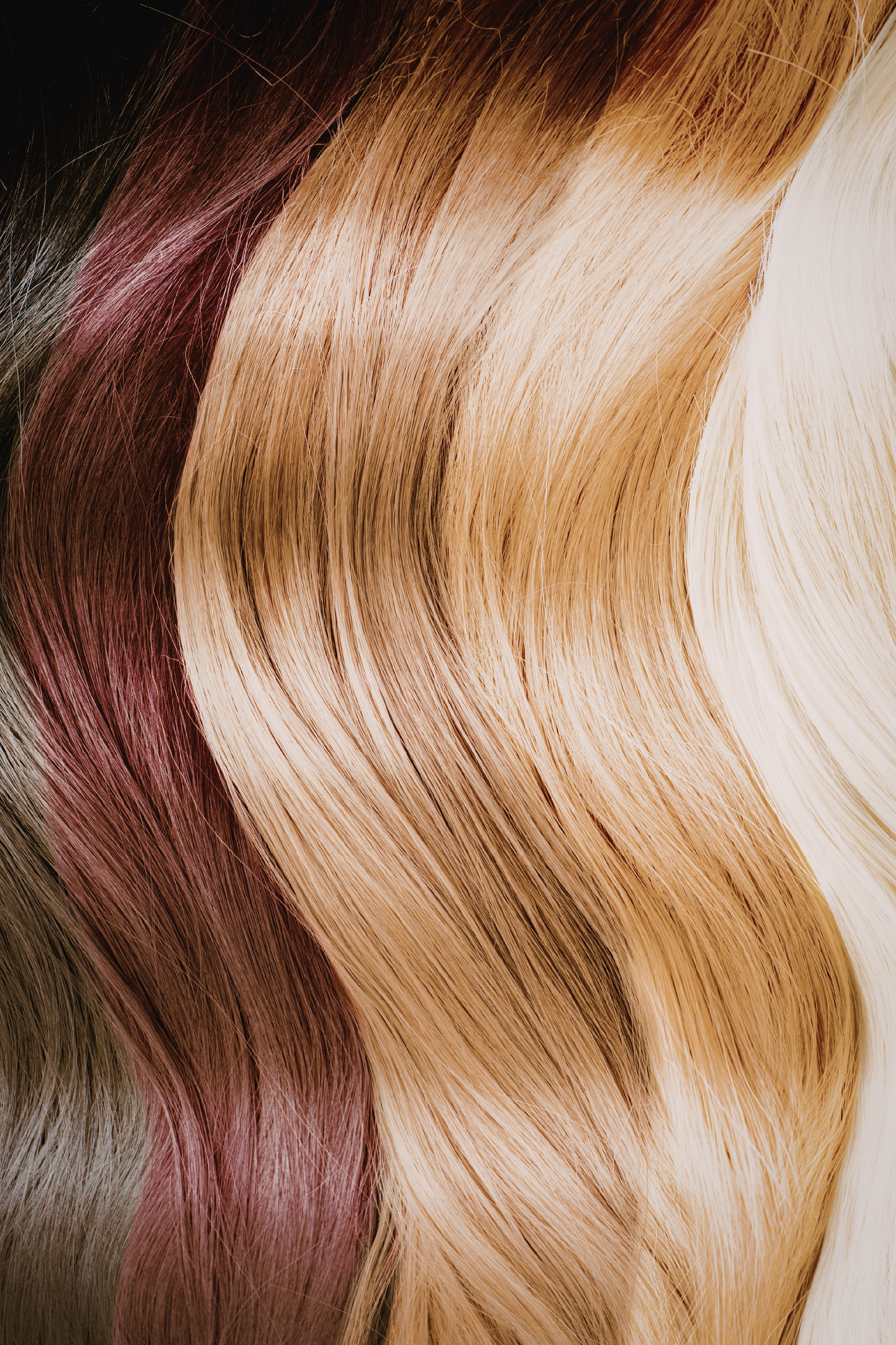 Nahaufnahme von gefärbtem, langem, glattem Haar in verschiedenen Farben wie Braun, Blond und Weiß, welche in sanften Wellen fallen.