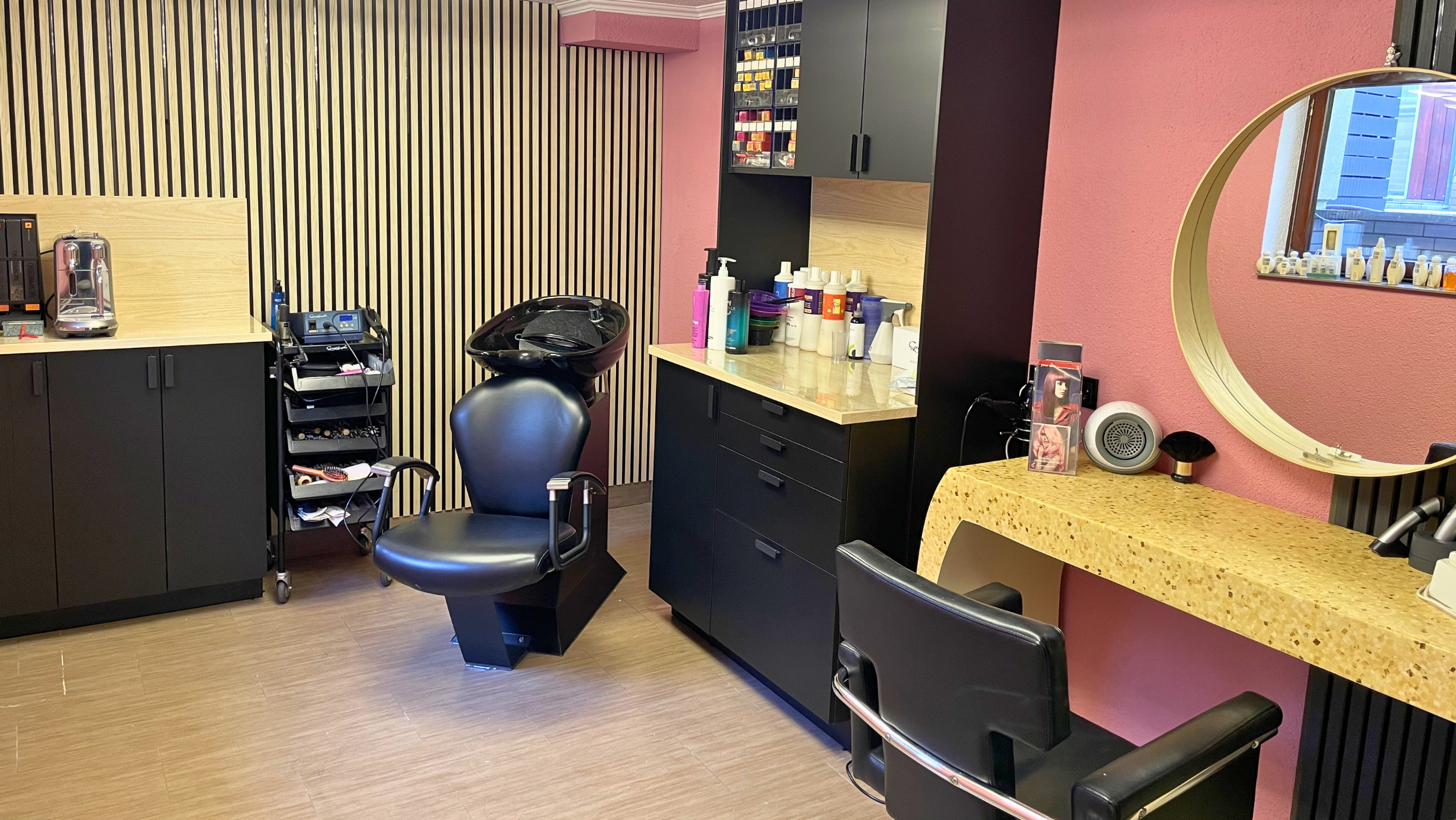 Friseursalon Maya mit schwarzen Schränken, einem Waschbecken, einem Spiegel, einem schwarzen Stuhl, Pflegeprodukten, Handtüchern und Kosmetikartikeln.