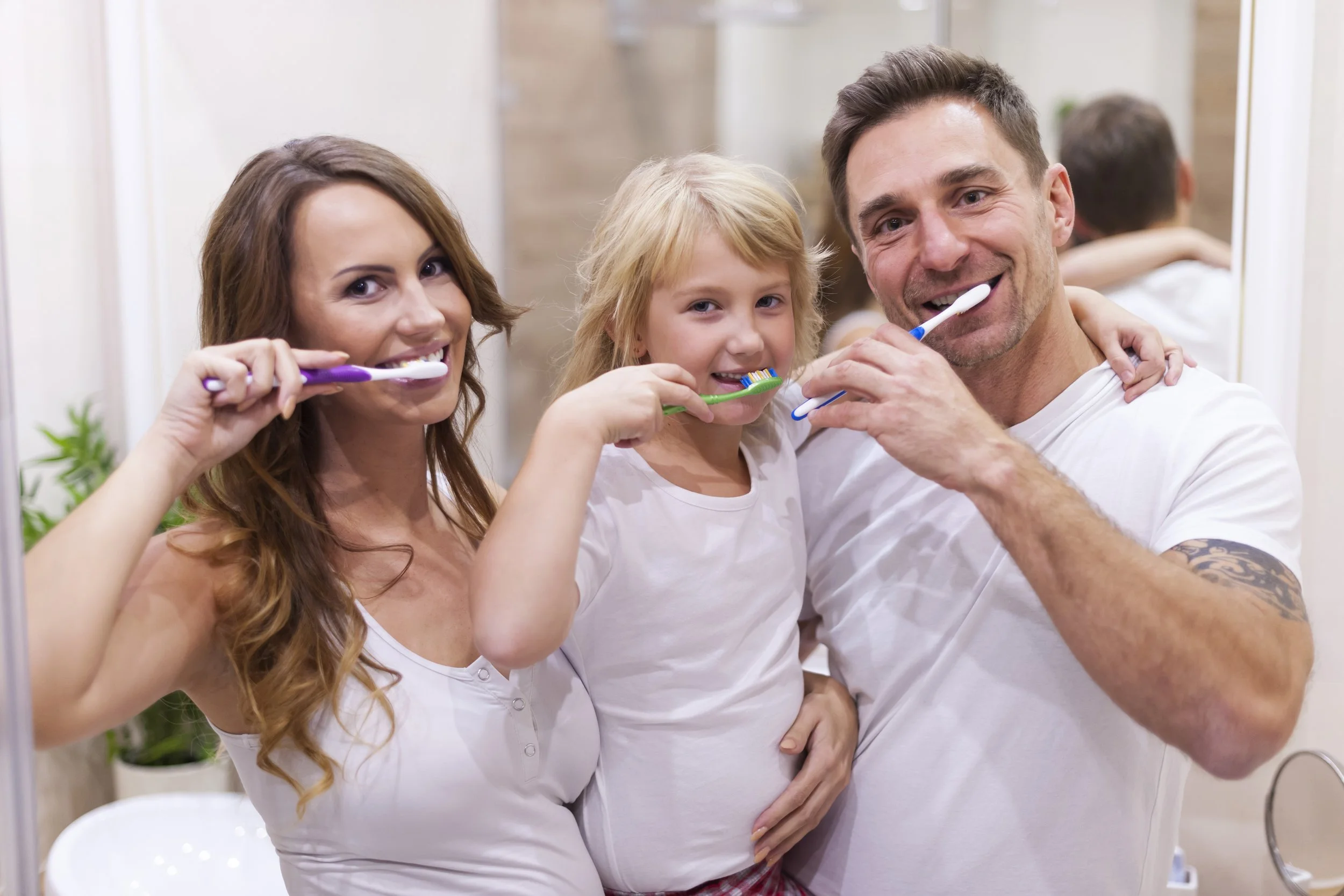 A family of three brushing their teeth together in a bathroom mirror, smiling happily.