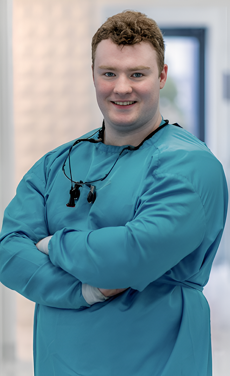 A young male healthcare professional in blue scrubs standing with his arms crossed in a medical facility.