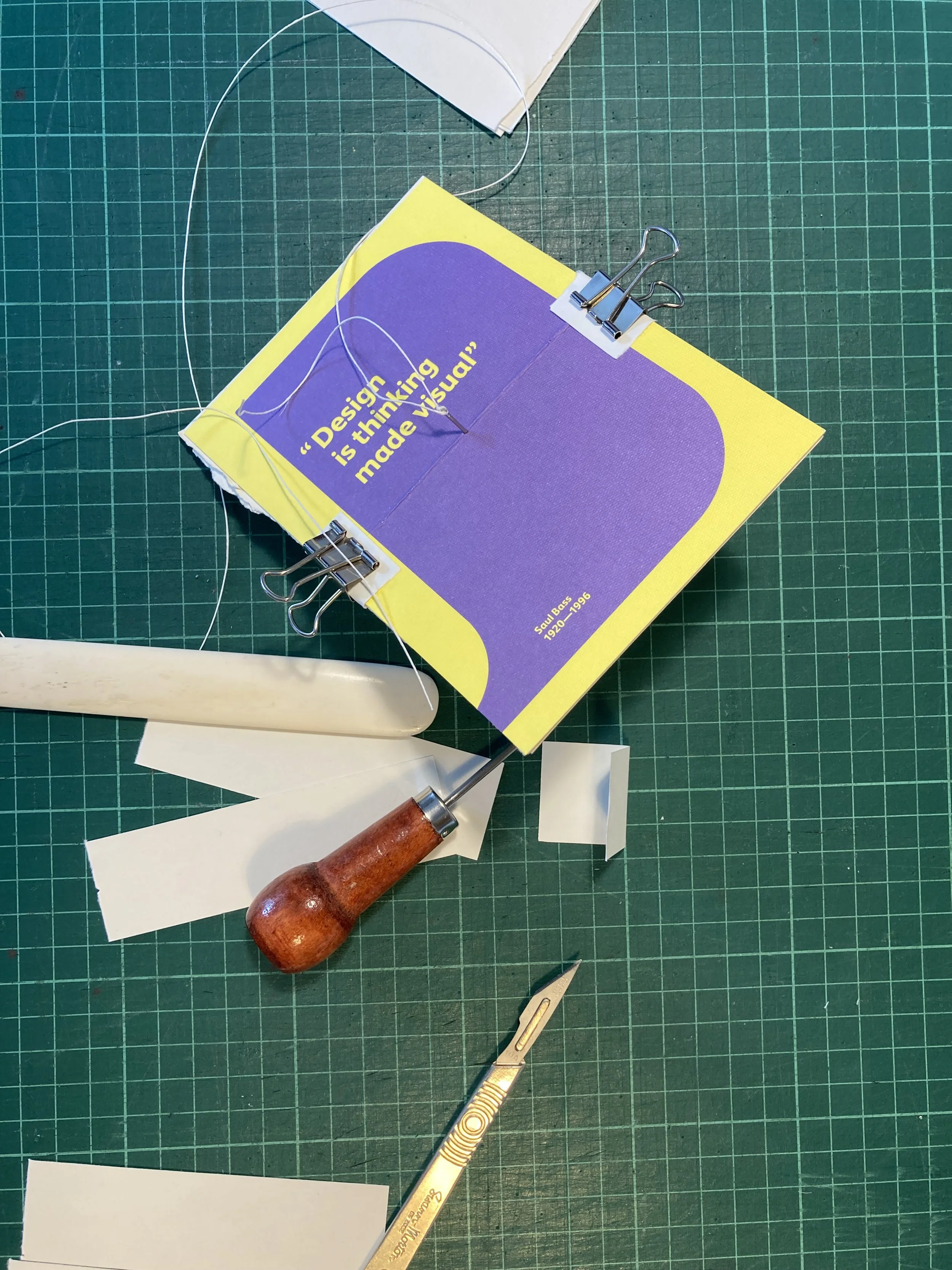 A green cutting mat with a yellow and purple booklet that has the quote "Design is thinking made visual" pinned with metal clips. There is a small section of white paper, a wooden handle tattoo needle, a gold utility knife, and small paper pieces scattered around.