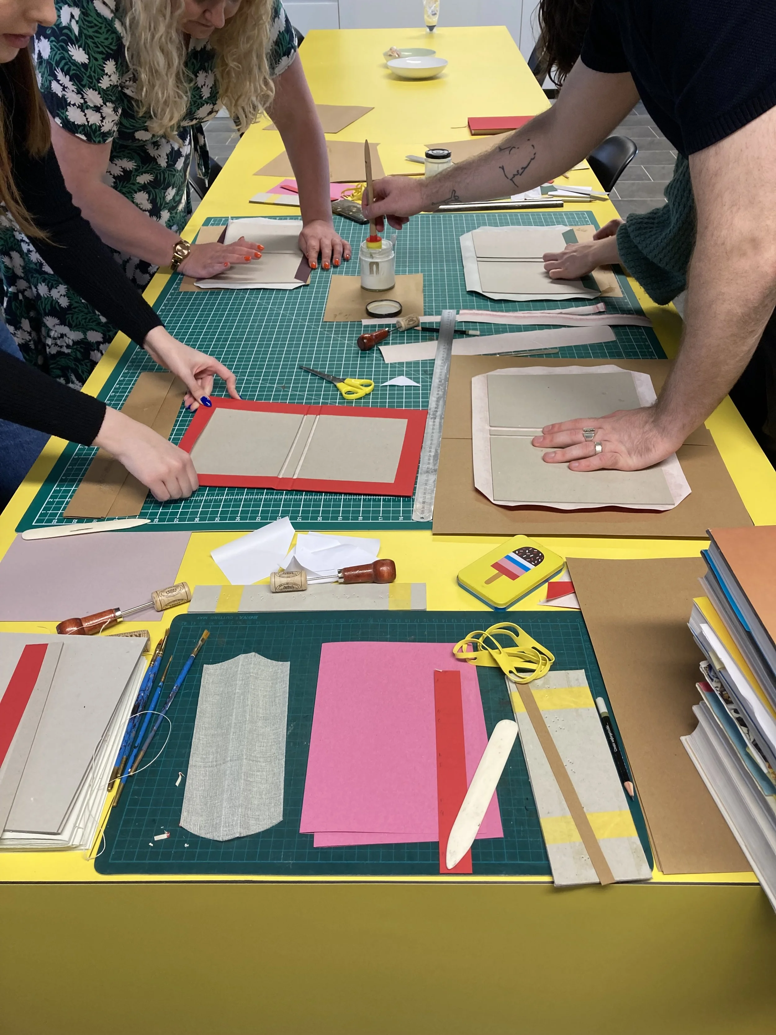 People gathered around a yellow table working with paper, fabric, and crafting tools, possibly in a workshop setting.