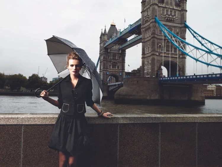 A woman in black dress holding a black and white umbrella stands near the River Thames with Tower Bridge in London in the background on a cloudy day.