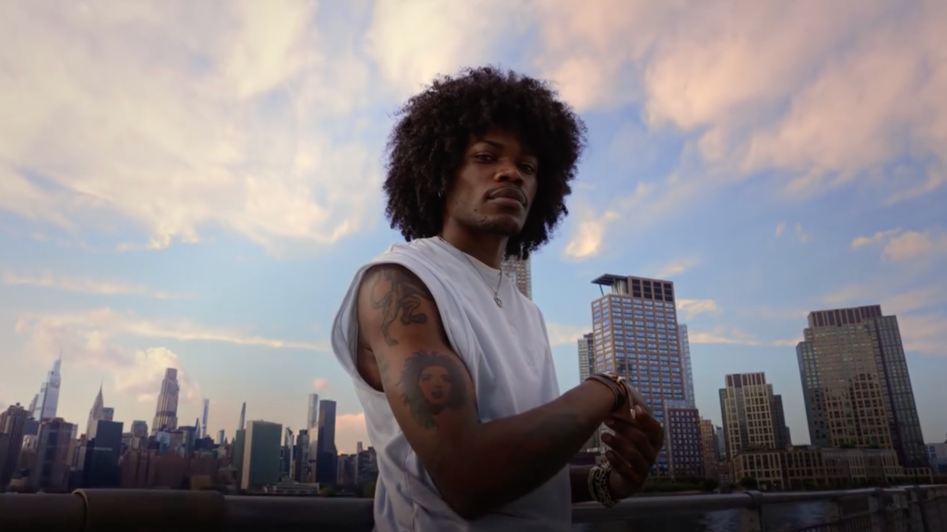 A man with curly hair and tattoos on his arm stands on a rooftop with a city skyline in the background during sunset or dusk.