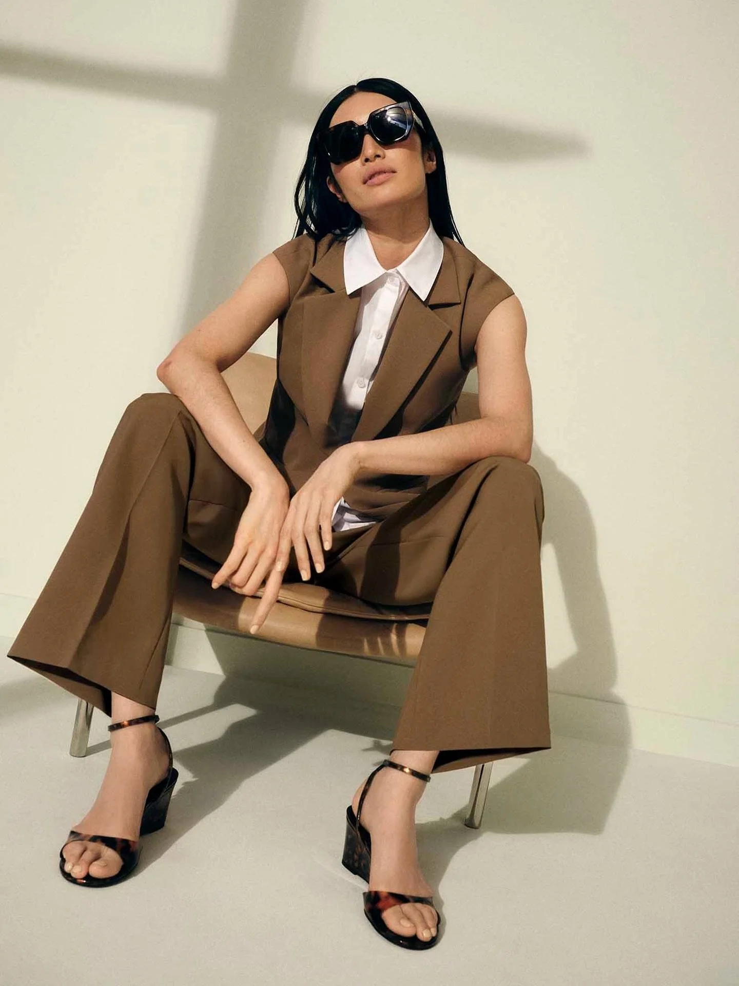Woman wearing black sunglasses, a sleeveless brown vest, a white shirt, brown wide-leg pants, and black high-heeled sandals, sitting on a beige chair against a plain light-colored background.