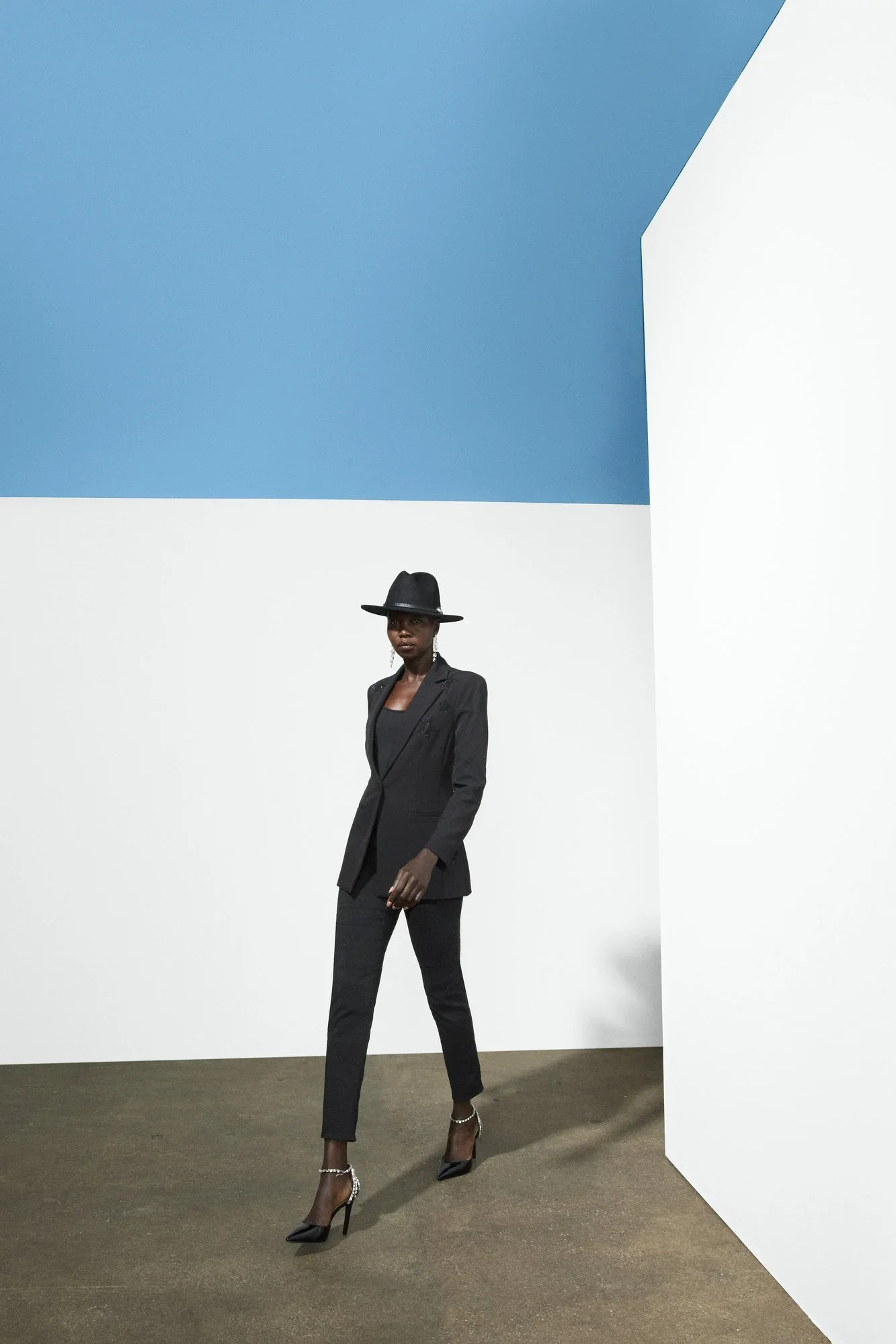 A woman dressed in a black suit, wearing high heels and a black wide-brim hat, standing against a modern abstract background with blue and white walls and a brown carpeted floor.