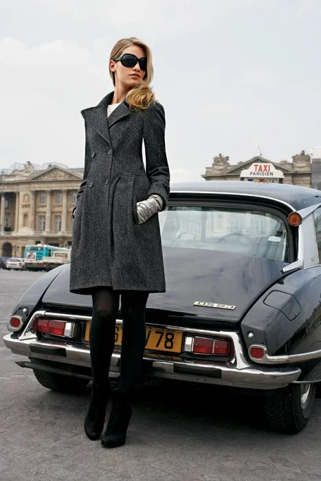 A woman in a long gray coat, sunglasses, and black high heels standing next to a vintage black car on a city street, with historic buildings and a taxi sign in the background.