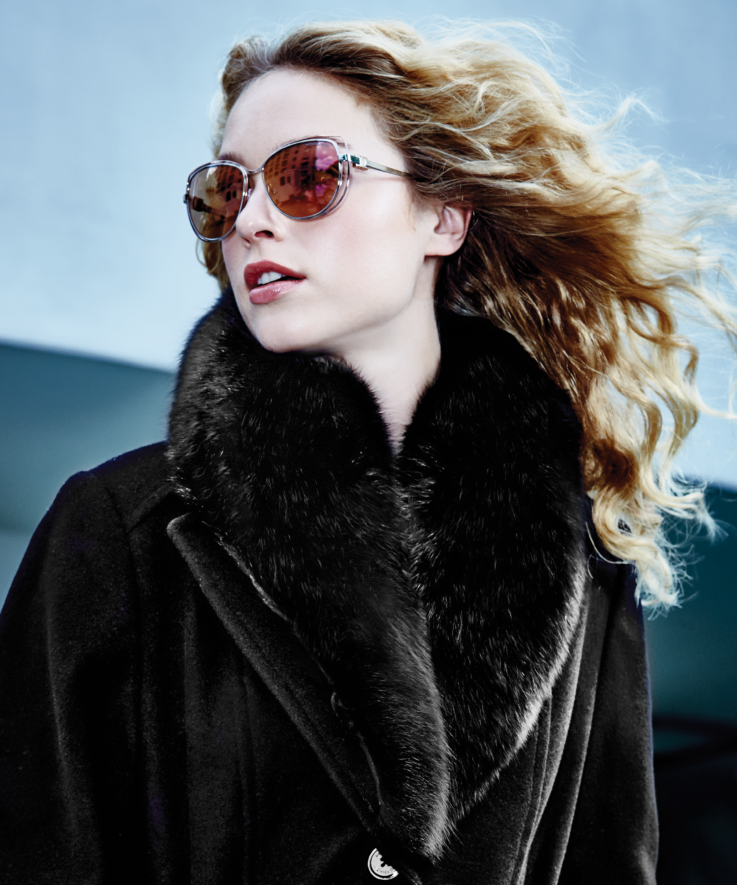 Close-up of a woman with curly blonde hair wearing reflective sunglasses and a black fur coat against a blue background.