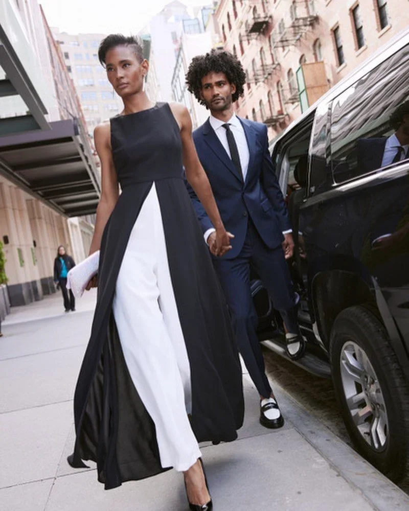 A stylish woman in a black and white gown walks past a man in a navy suit on a city sidewalk, holding hands. There is a black car parked nearby and buildings in the background.