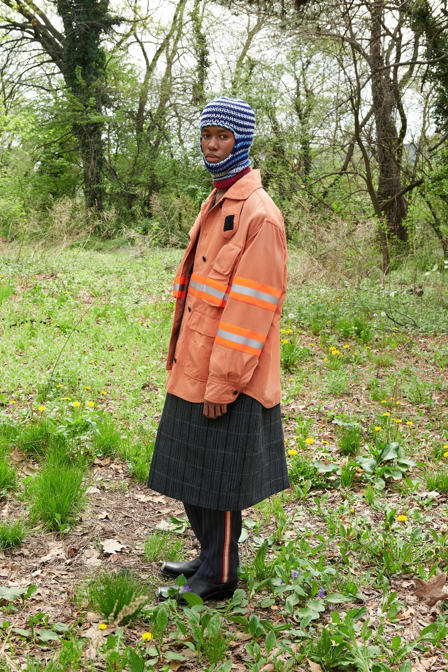 A person standing outdoors in a forested area with a light brown jacket with reflective stripes, a chequered skirt, and a blue and white knit balaclava.