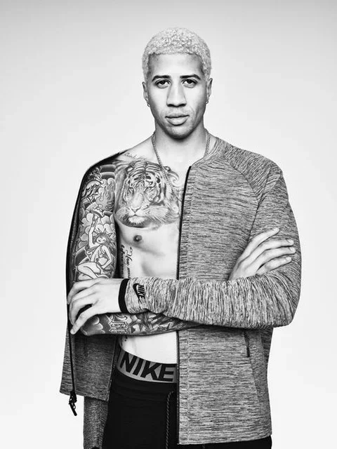 Black and white photo of a young man with bleached blonde hair, tattoos on his chest and right arm, wearing a zip-up hoodie and Nike underwear, standing with arms crossed against a plain background.
