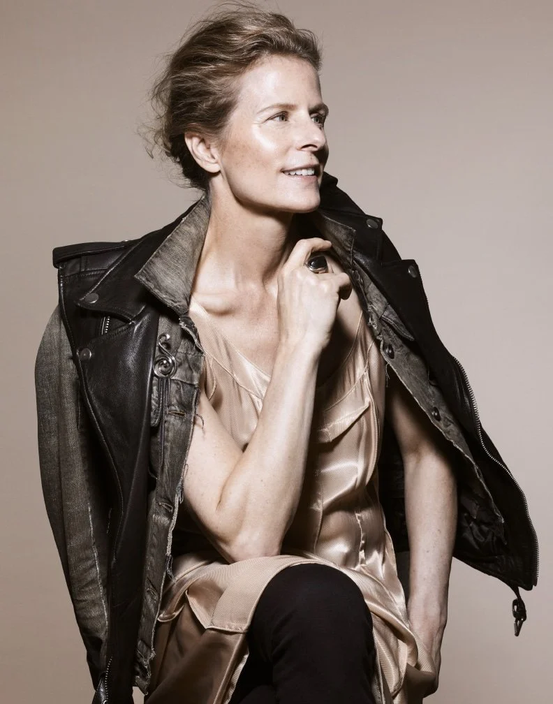 A smiling woman with short, wavy blonde hair wearing a beige satin dress and a black leather jacket, sitting against a plain background.