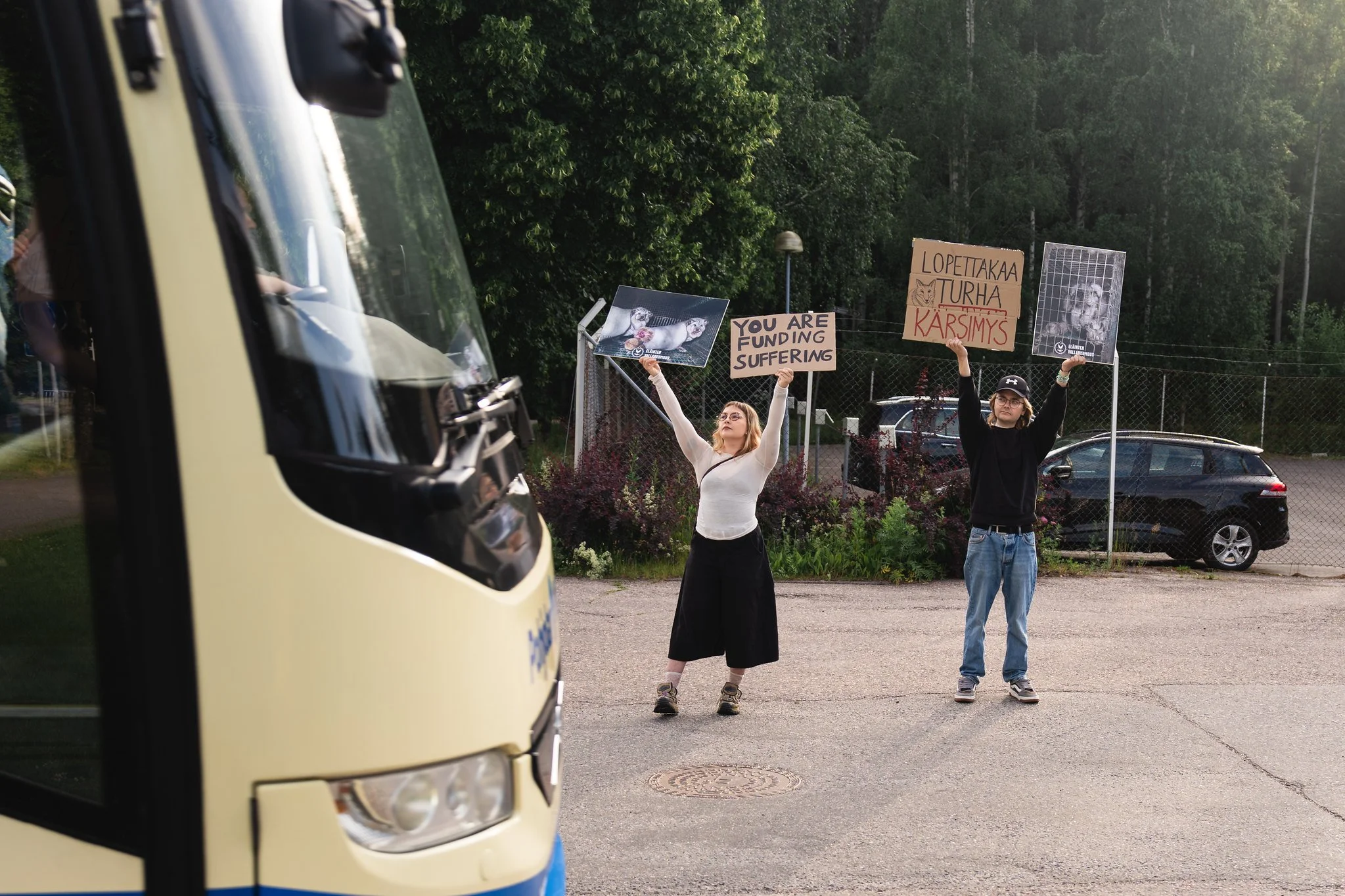 Kaksi ihmistä seisoo kadulla protestin merkeissä, kädessään kylttejä ja valokuvia eläinten suojelusta. Taustalla puita ja autoja, näkyvissä myös rautatie.