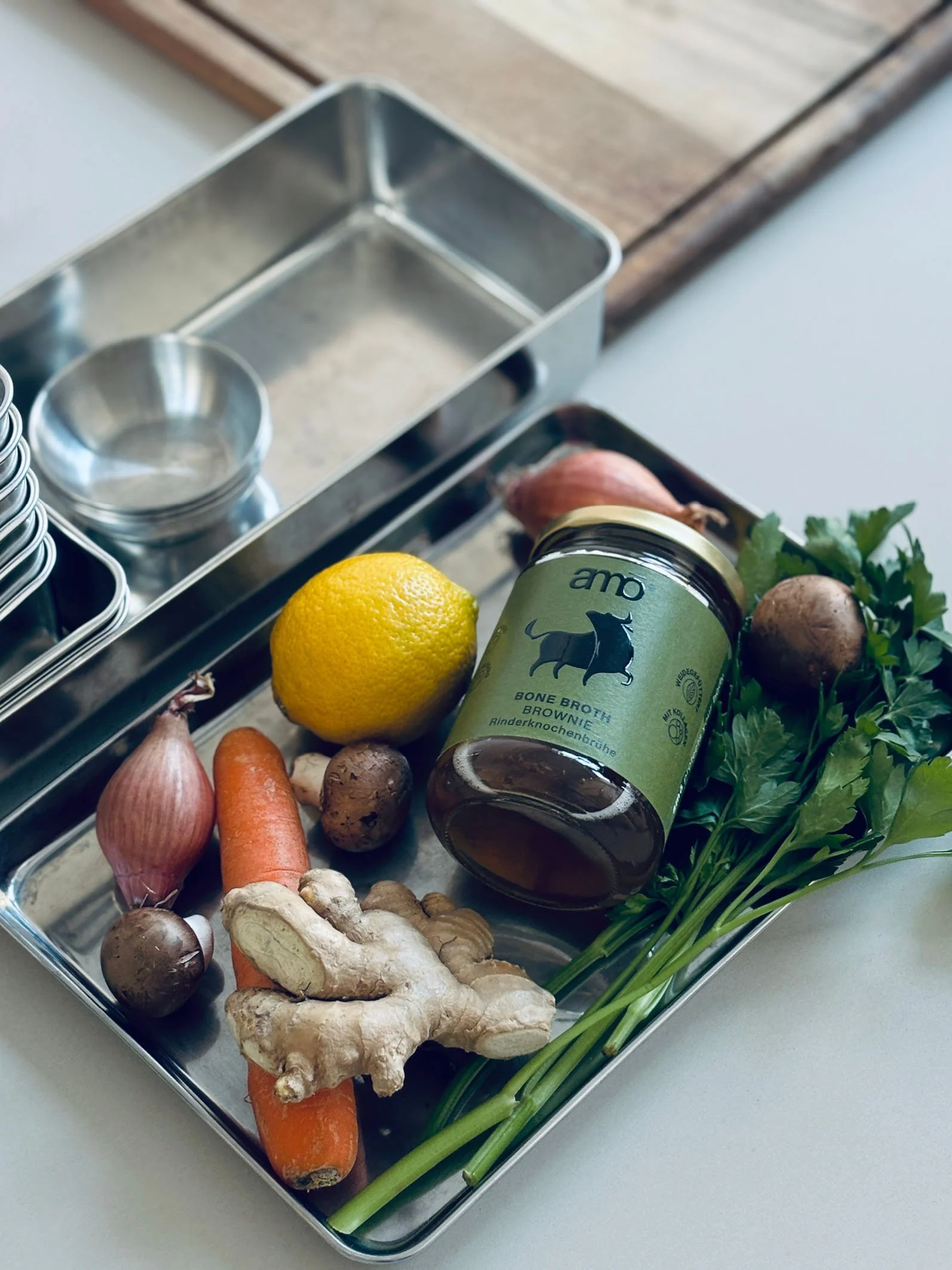 Ein Metalltablett mit frischen Zutaten, darunter eine Zitrone, Karotten, Ingwer, Knoblauch, Shiitake-Pilze, Petersilie, ein Glas mit Knochenbrühe, Zwiebel und Frühlingszwiebeln.