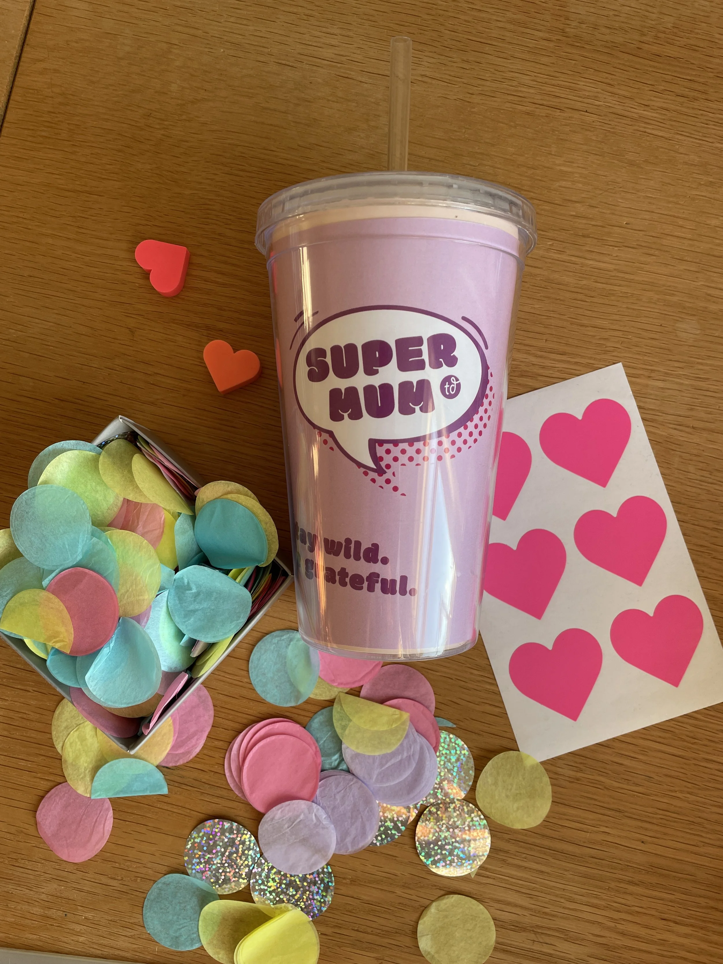 Ein durchsichtiger Becher mit rosa Beschriftung, umgeben von bunten Papierkreisen und herzförmigen Aufklebern auf einem Holztisch.