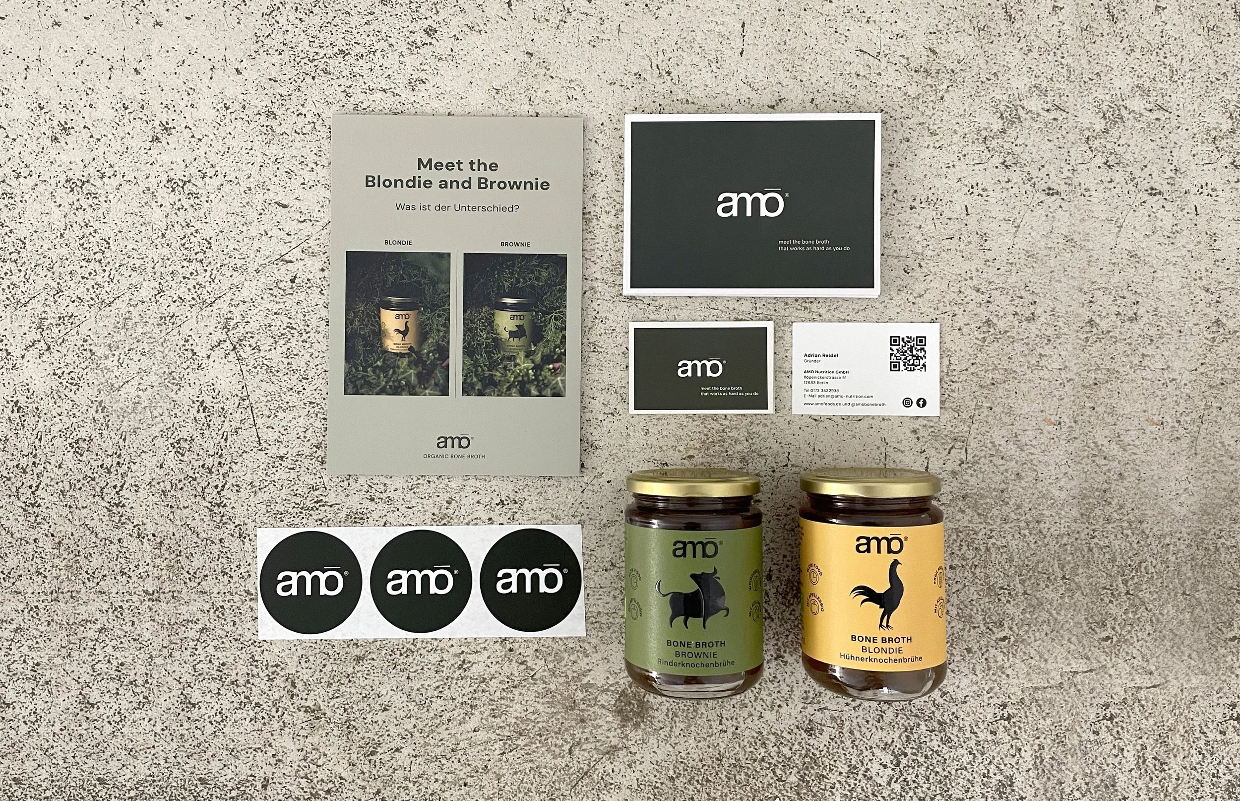 Produktbild mit verschiedenen Gläsern und Verpackungen von bone broth mit Markenlogo amö, einem Flyer mit Vergleich zwischen Blondie und Brownie und weiteren Broschüren sowie Aufklebern mit dem Logo amö auf einer helle, steinartige Oberfläche.