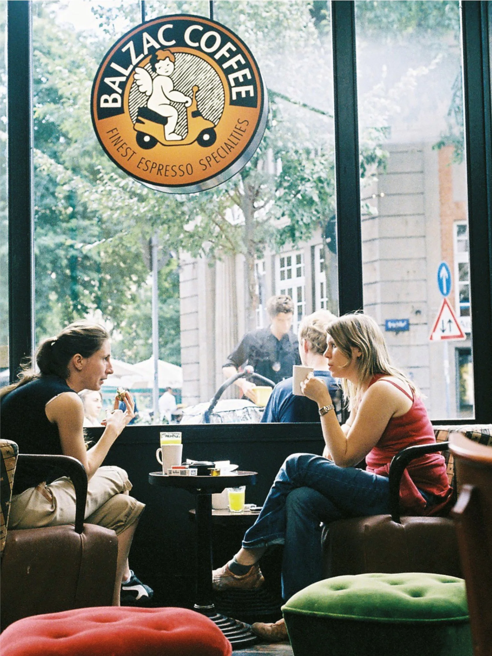Menschen sitzen in einem Café und trinken Kaffee, im Hintergrund große Fenster mit Blick auf die Straße und die Stadt. Das Logo des Cafés „Balzac Coffee“ ist an der Scheibe zu sehen.