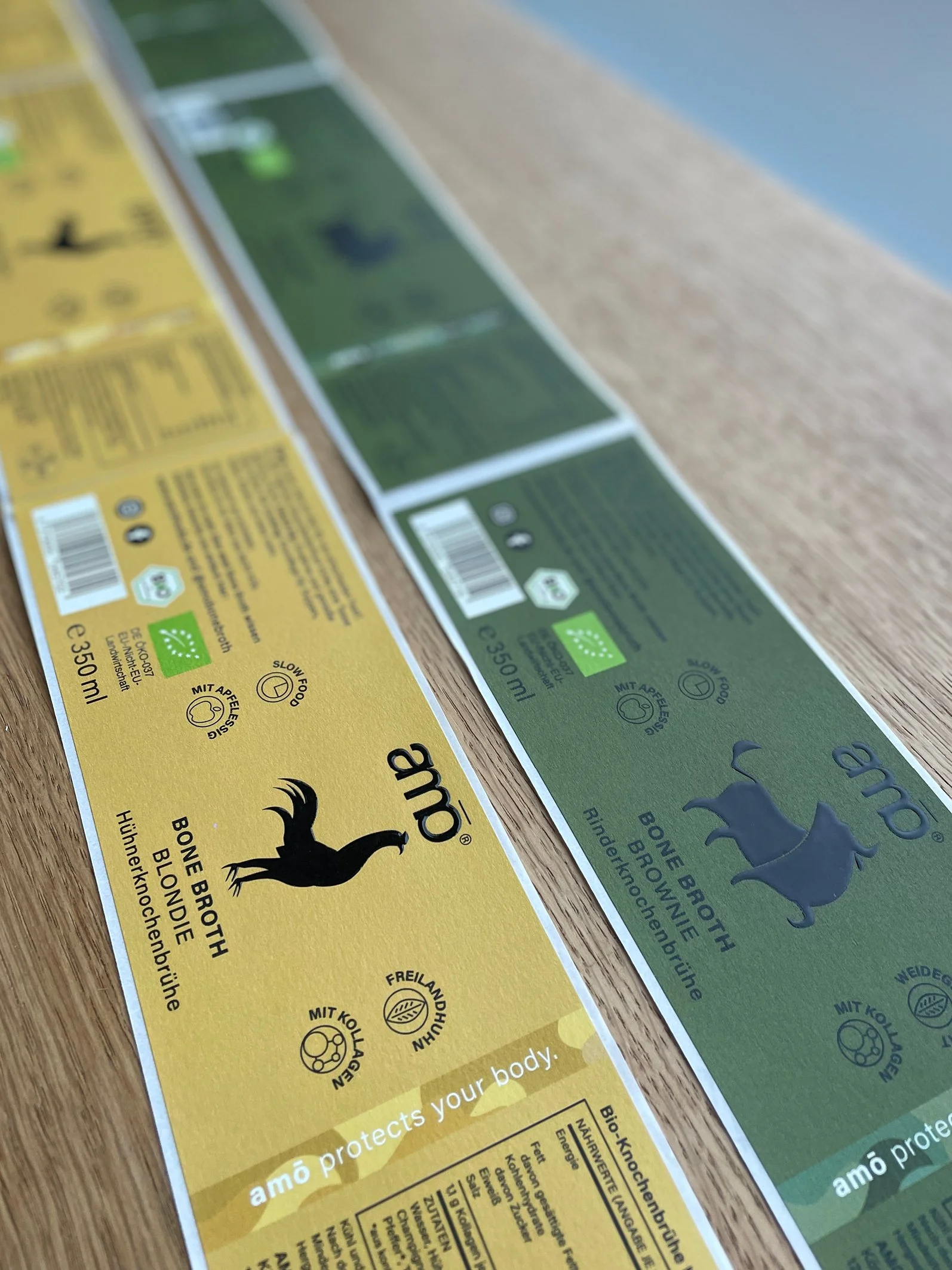 Diese Abbildung zeigt mehrere Etiketten für amö Protekt, mit einer gelben und grünen Verpackung, die auf einer holzfarbenen Oberfläche liegen.