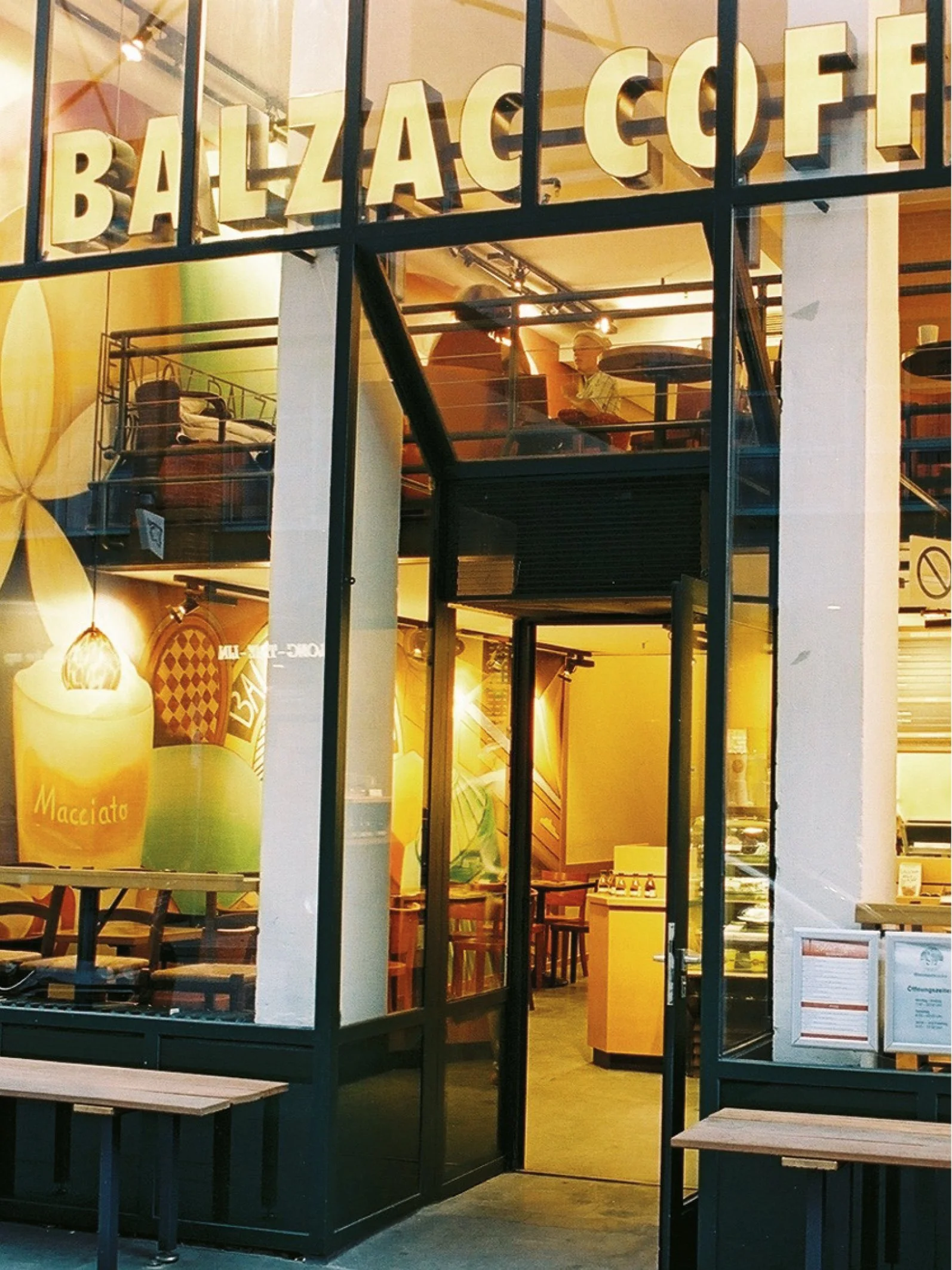 Ansicht eines hell erleuchteten Cafés mit großen Fenstern, Innenbereich mit Tischen und Stühlen, personale und dekorative Elemente sichtbar.