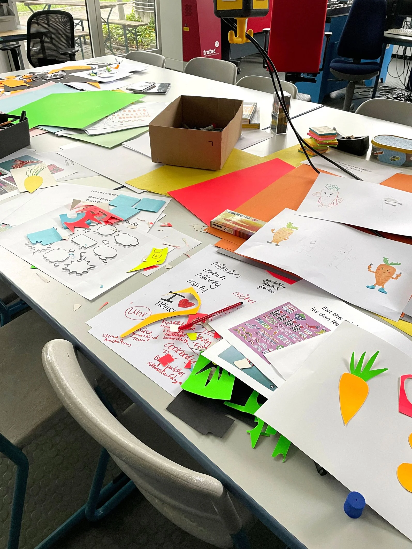 Lehrbeauftragung der Hochschule München und ein Tisch mit verschiedenen Bastelmaterialien, Papier, Stiften und Papierausschnitten