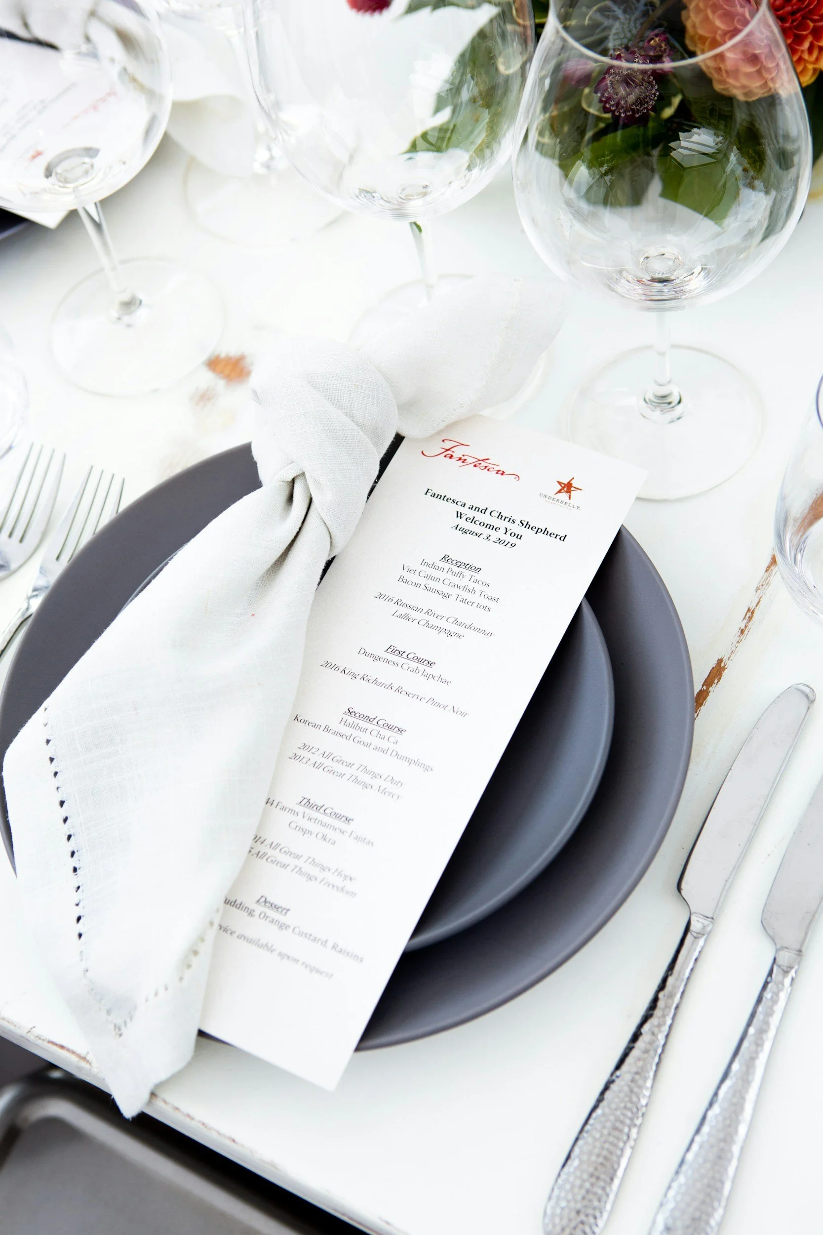 Table setting with empty wine glasses, a white napkin tied with a knot, a printed menu, a gray plate, and silverware on a white surface.