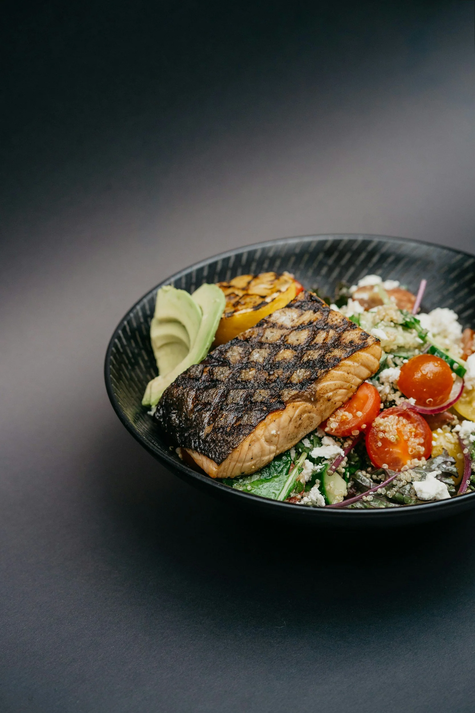 A black bowl with grilled salmon, slices of avocado, roasted lemon, cherry tomatoes, and a colorful vegetable salad on a black surface.