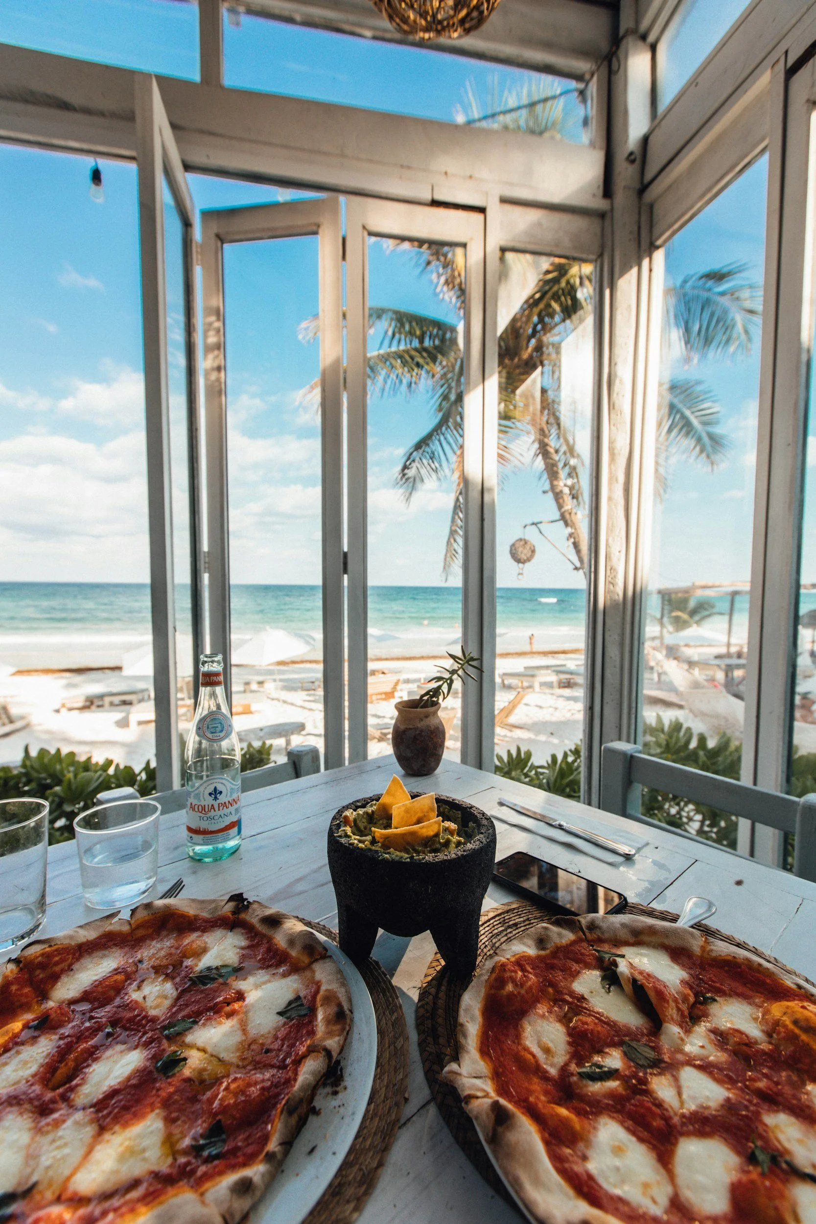 A table with two large cheese and tomato pizzas, a glass bottle of water, and a glass of water, all set outdoors with a view of the beach, ocean, and palm trees through glass windows.
