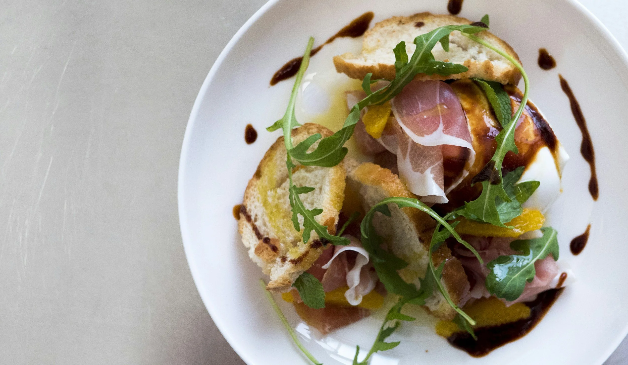 A white bowl with a bruschetta topped with arugula, prosciutto, cheese, and a drizzle of balsamic glaze.