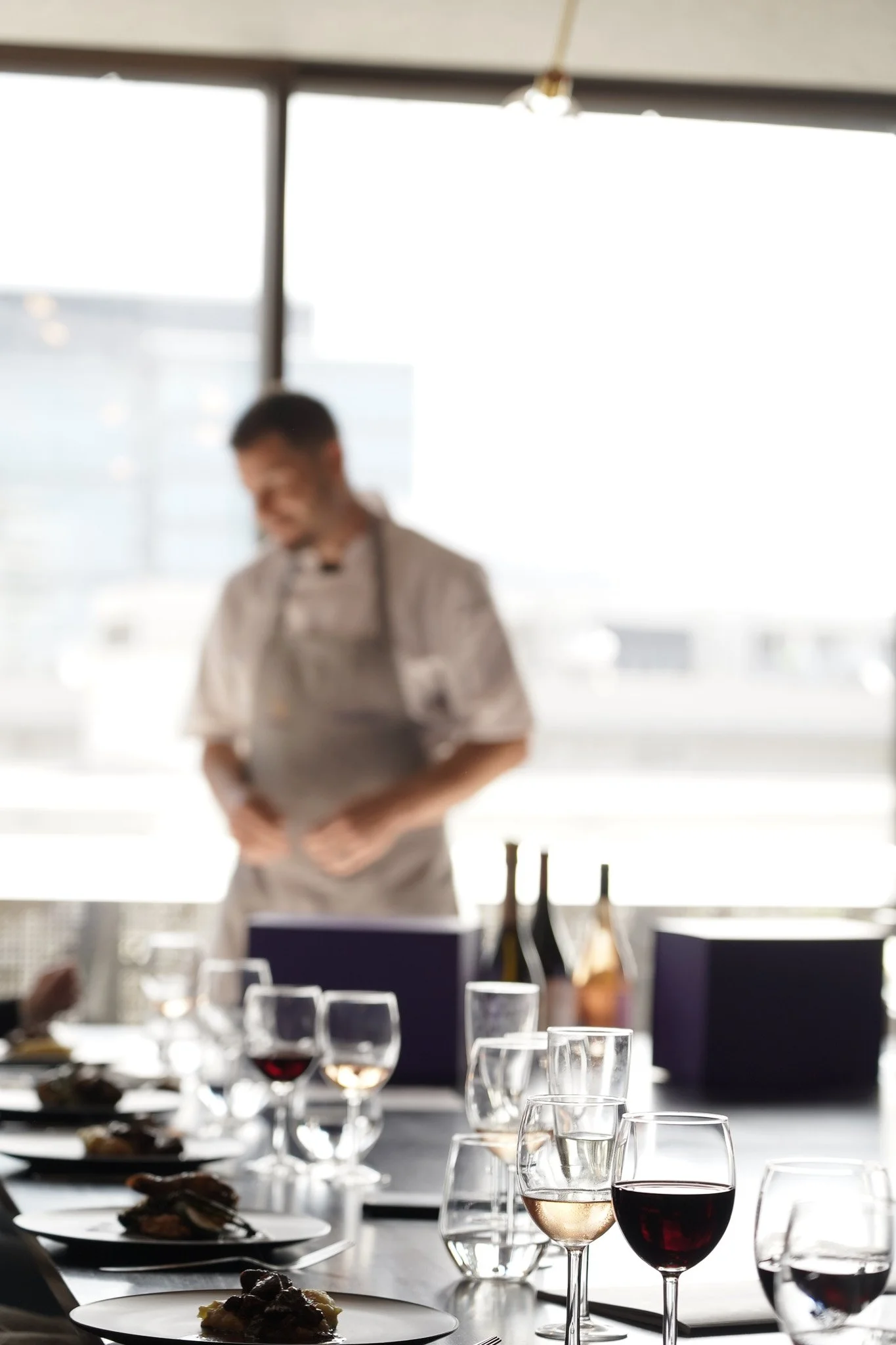 A person wearing a chef's apron smiling behind a table set with wine glasses and plates of food in a bright, modern dining area with large windows.