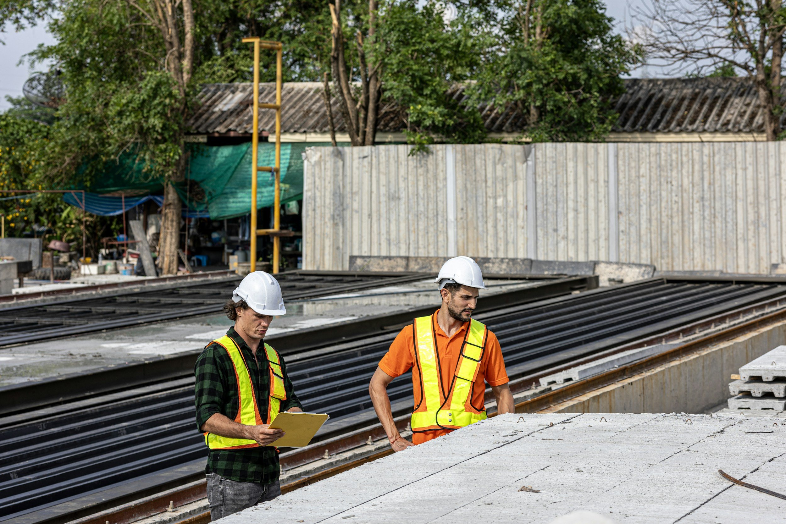 Twee bouwvakkers op een bouwplaats onderzoeken een betonnen vloer, met één die een clipboard vasthoudt en de ander die aantekeningen maakt, terwijl ze een zonsverduisteringsbril dragen.  Ze werken voor Vondr. Recruitment. Vondr jobs, vondr werk