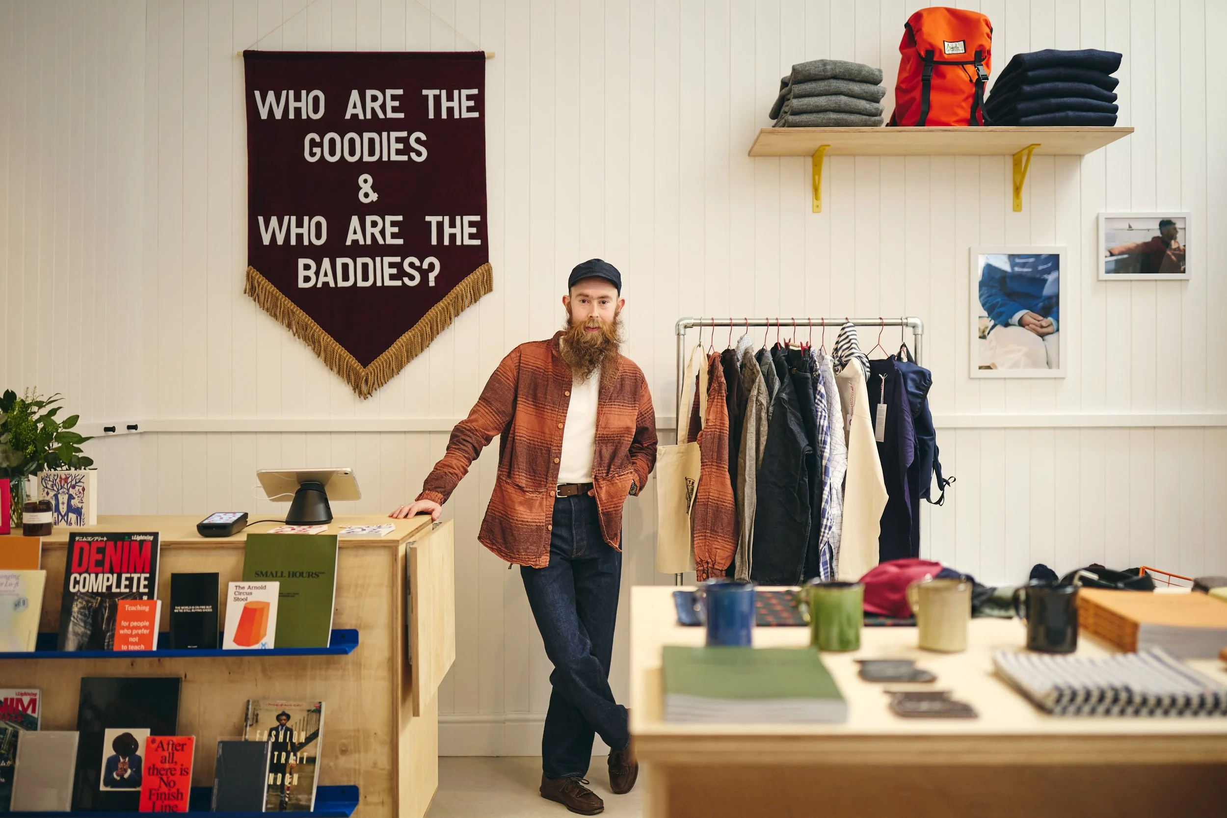 A man with a beard and wearing a cap, brown plaid shirt, and dark pants, standing behind a retail counter inside a clothing store. Clothes are hanging on a rack next to him, and shelves with folded shirts and a red backpack are above. A maroon banner with white text asks, "Who are the goodies & who are the baddies?" on a white wall behind him.