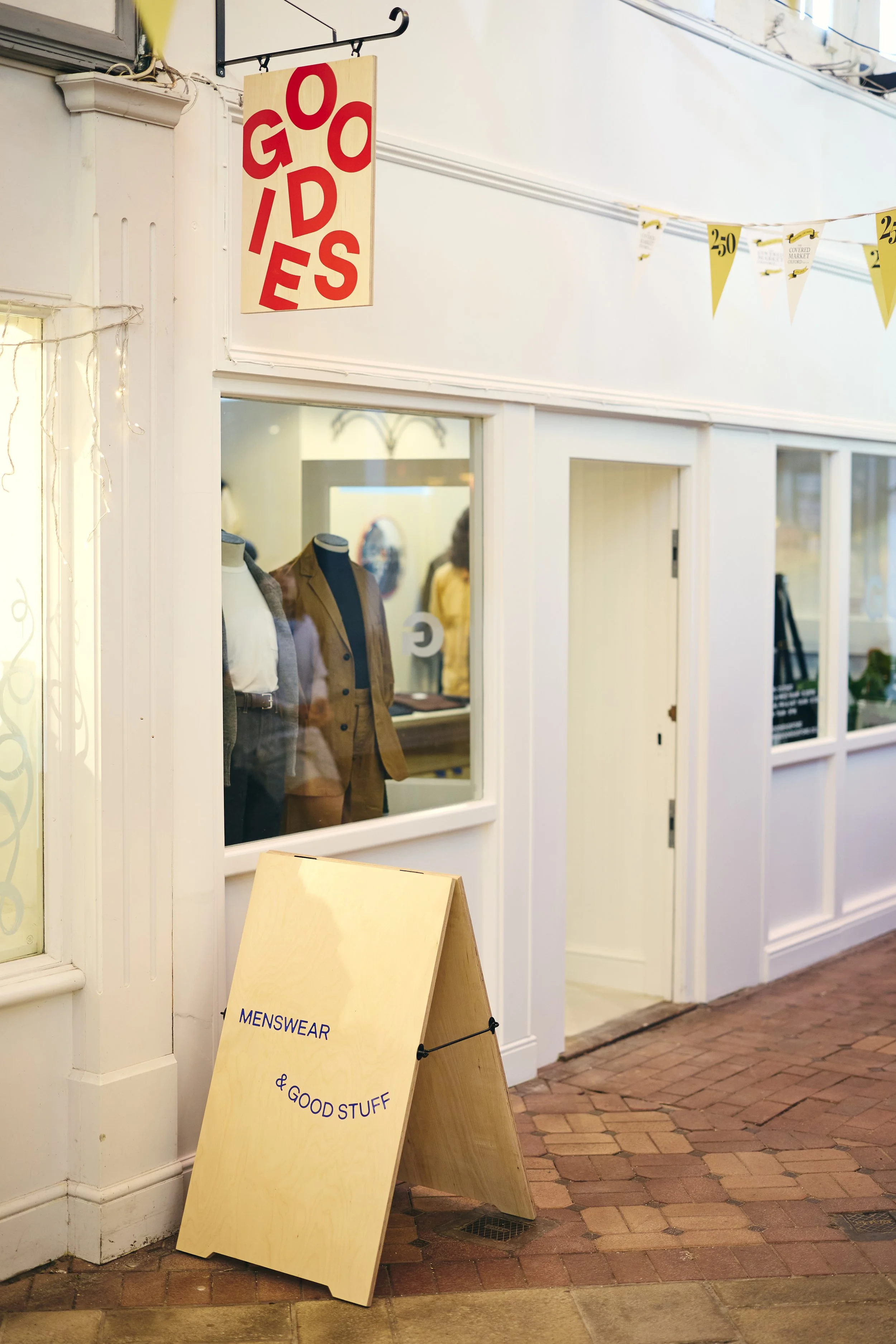 Street view of a menswear store with a large wooden A-frame sign on brick sidewalk that reads 'MENSWEAR & GOOD STUFF.' Window displays mannequins dressed in men's suits and jackets. A hanging sign above the window reads 'GOODS' with the letters arranged vertically.