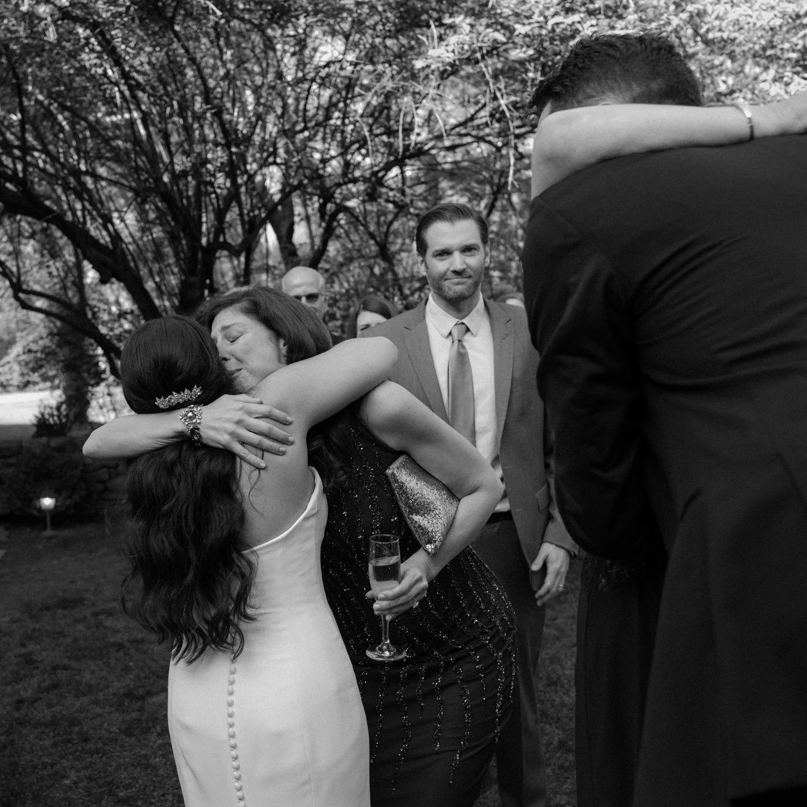 People hugging at a wedding reception outdoors, some holding drinks, with trees in the background.