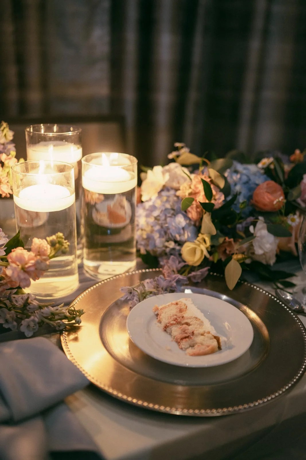 Elegant table setting with three lit candles, a floral arrangement, a tray with a partially eaten pastry on a plate, and a dark background.
