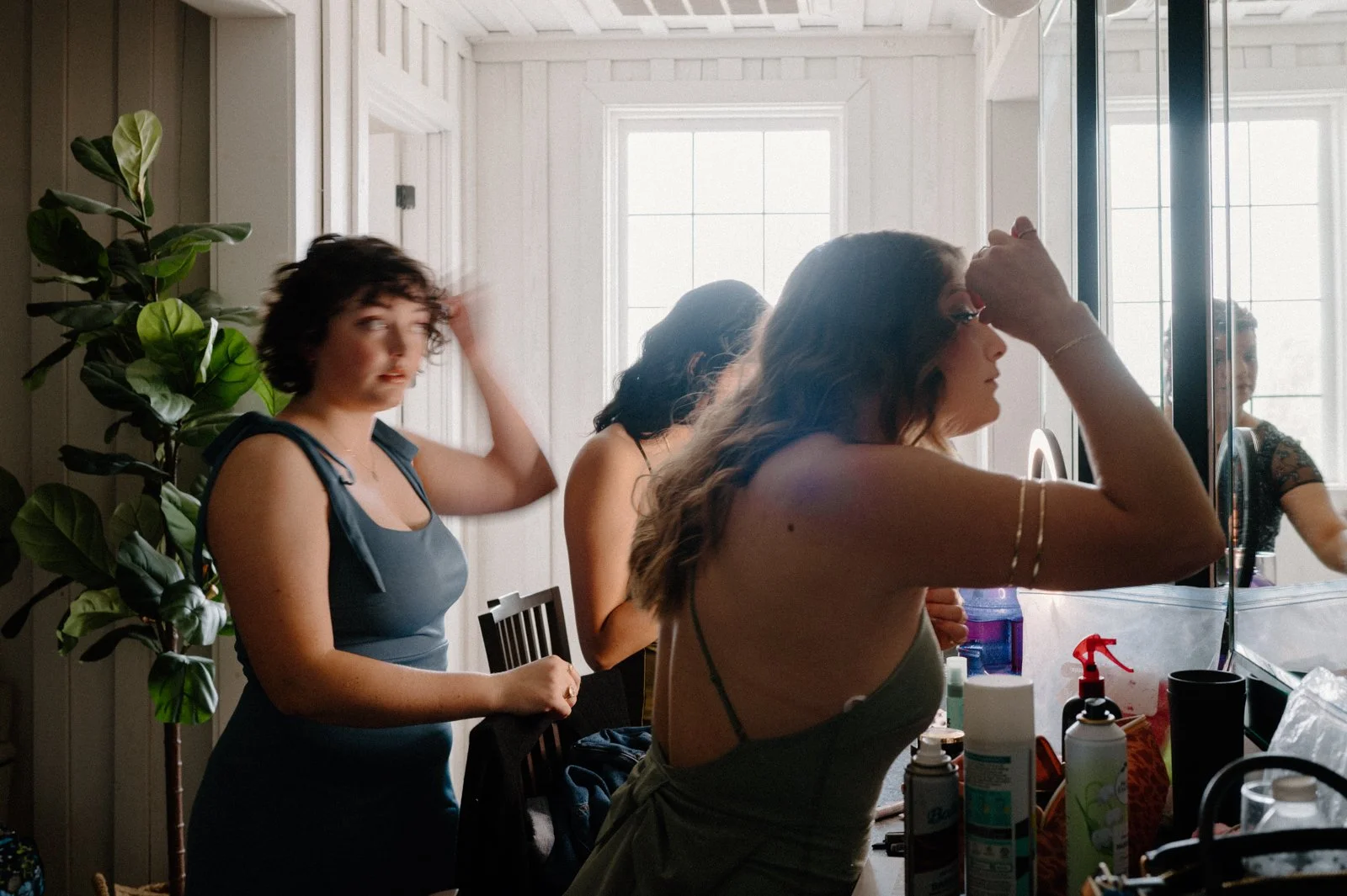 Three women in a well-lit room with large windows, one is standing and fixing her hair, another is seen from behind, and the third woman is looking into a mirror. The space appears to be a bathroom or dressing area with various personal care products