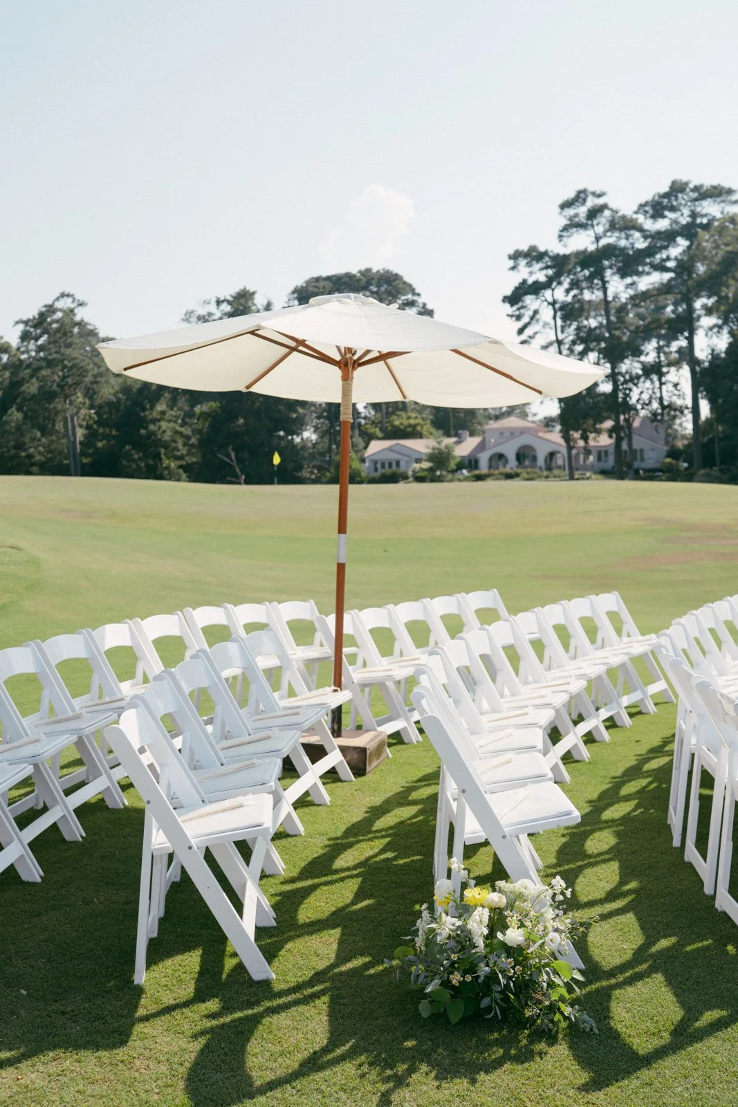 Outdoor wedding seating arrangement on a grassy field with white folding chairs, a large white umbrella, a flower arrangement, and a residential house in the background.