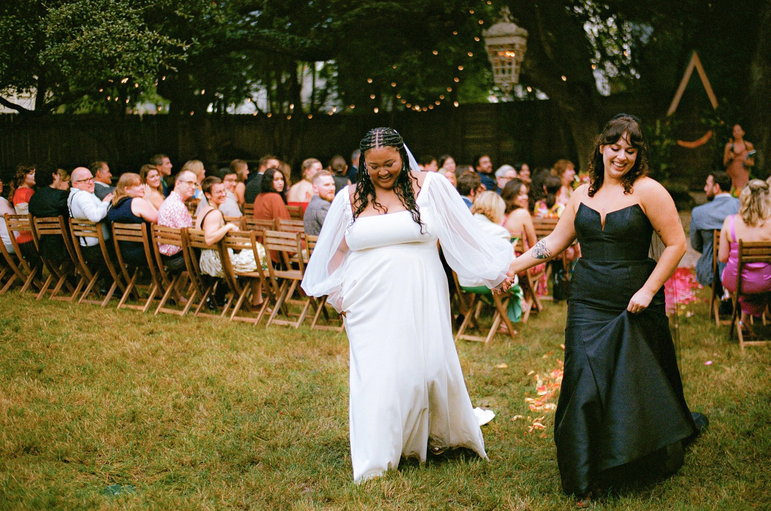 Two women dancing at an outdoor wedding ceremony, with many seated guests in the background. One woman is wearing a white dress with long sleeves and braided hair, and the other woman is in a black strapless dress with curly hair and tattoos, both sm