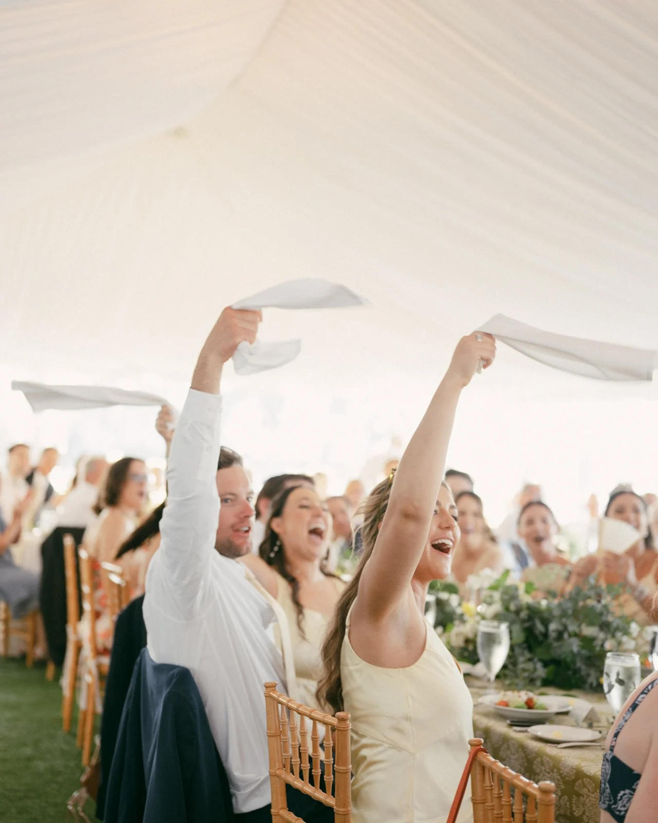 Crowd of people seated at tables during a celebration, with some raising white napkins in the air, smiling and cheering.