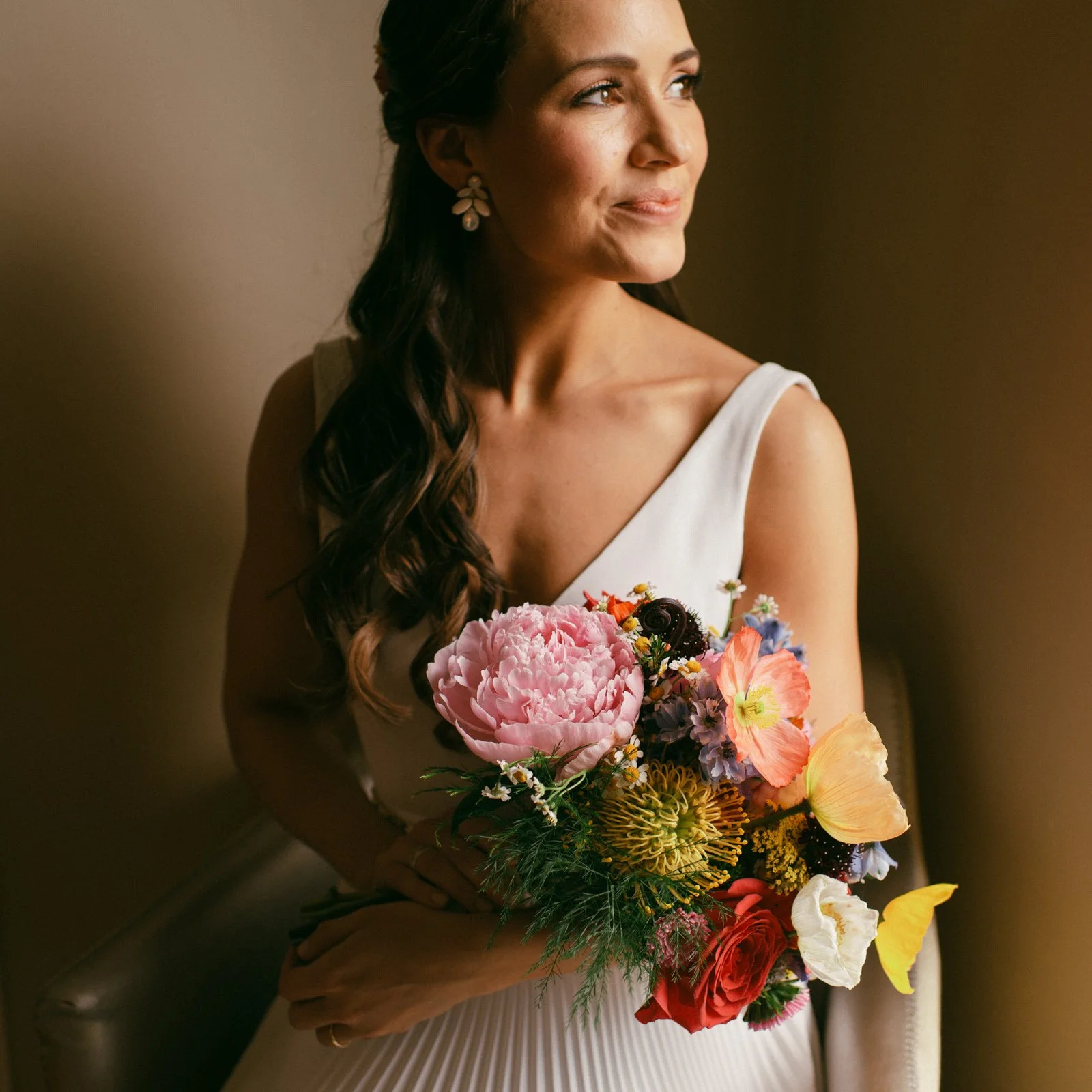 A woman with dark hair styled in loose curls, wearing a white sleeveless dress and floral earrings, holds a colorful bouquet of various flowers including pink, red, yellow, and purple blossoms, looking thoughtfully to the right.