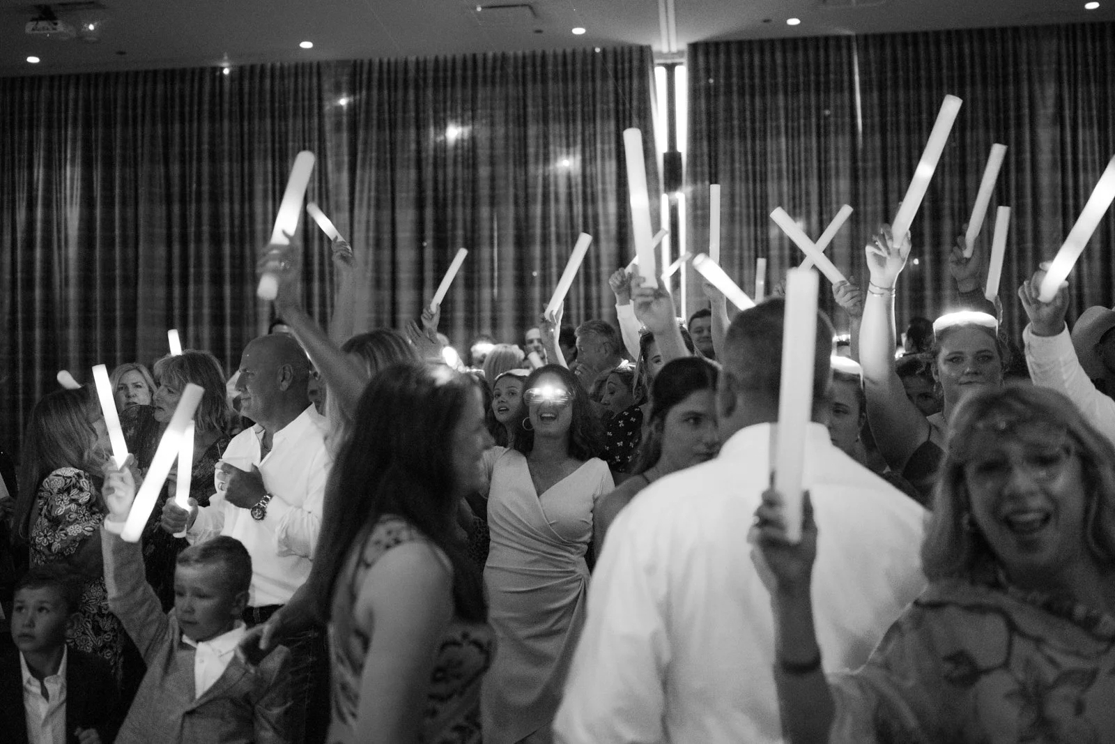 A large group of people at an indoor event holding illuminated light sticks, with some raising their light sticks in the air and others smiling and talking.