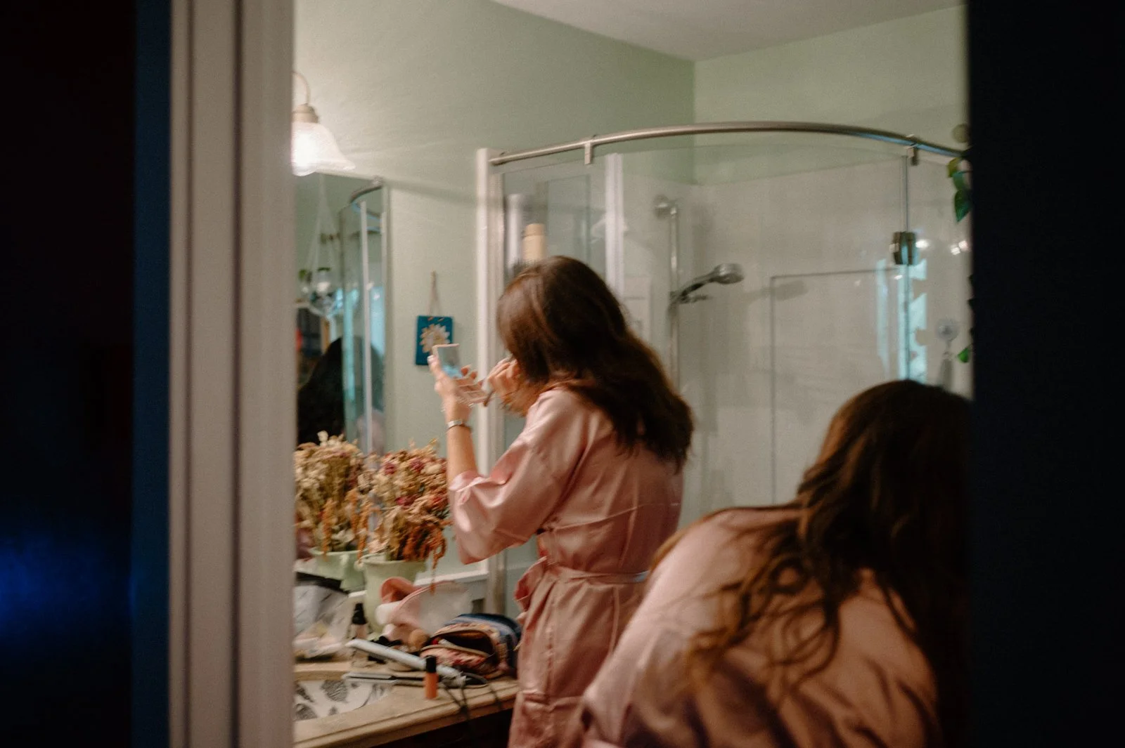 A woman with brown hair, wearing a pink satin robe, is standing in front of a bathroom mirror applying makeup or skincare. Another woman with long brown hair, also in a pink satin robe, is seated at the bathroom counter. The bathroom has a glass show