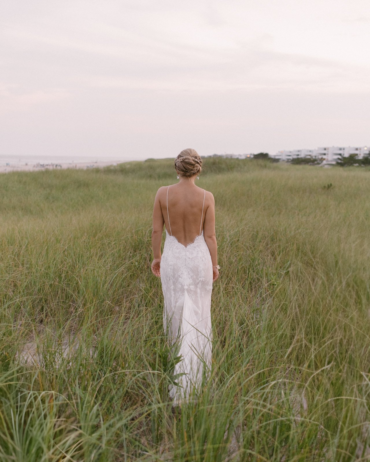 A bride in a white lace wedding dress with a low back walking through a grassy field at sunset, with a cloudy sky and modern buildings in the distance.