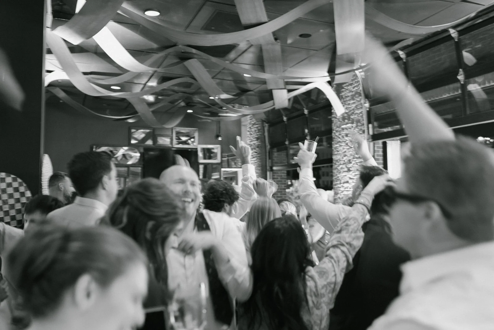 Group of people enjoying a celebration or party indoors with decorative streamers hanging from the ceiling, some raising their hands, and others smiling, in a lively setting with brick walls and framed pictures.