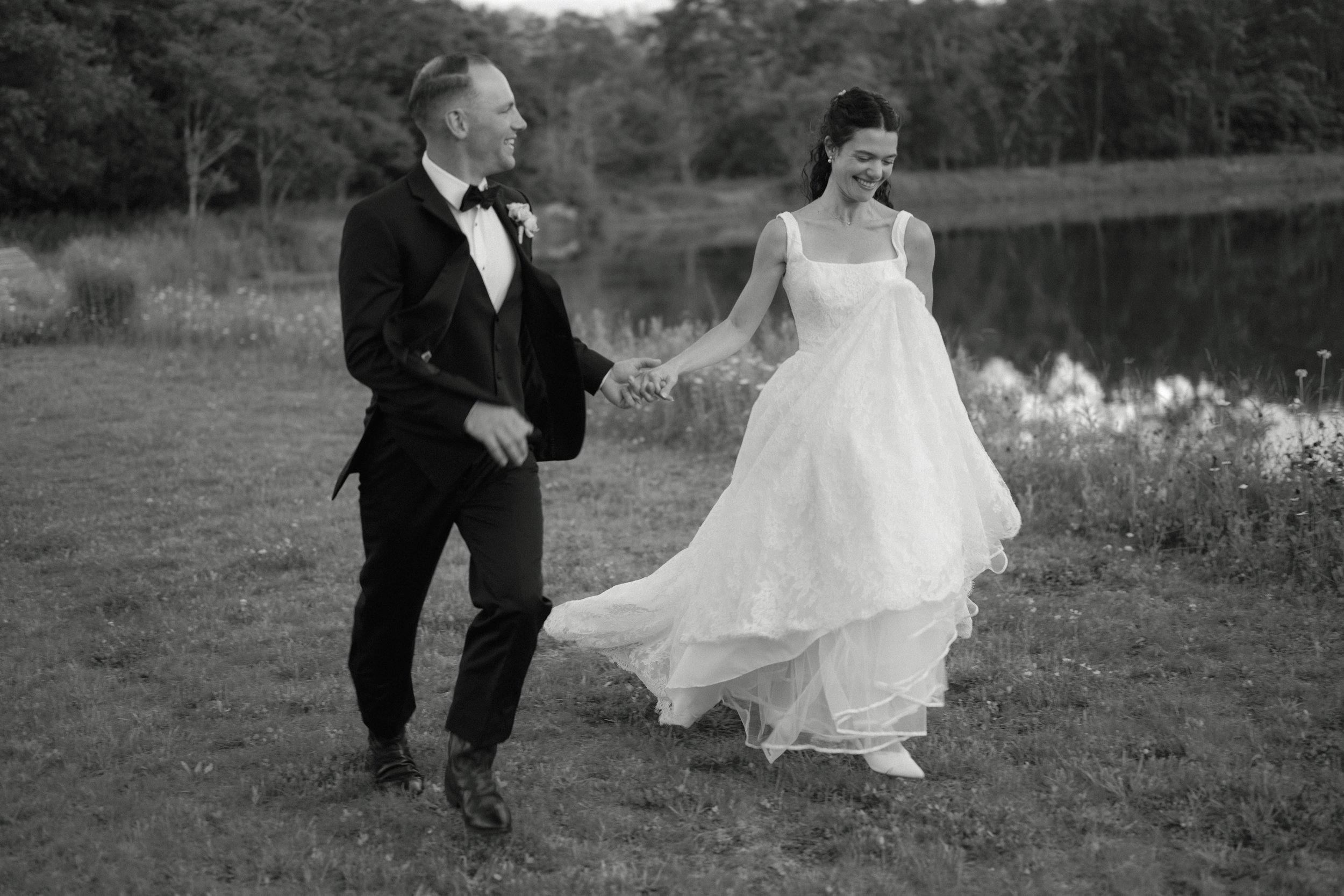 A black and white photograph of a bride and groom walking hand in hand outdoors near a lake, smiling and enjoying each other's company.