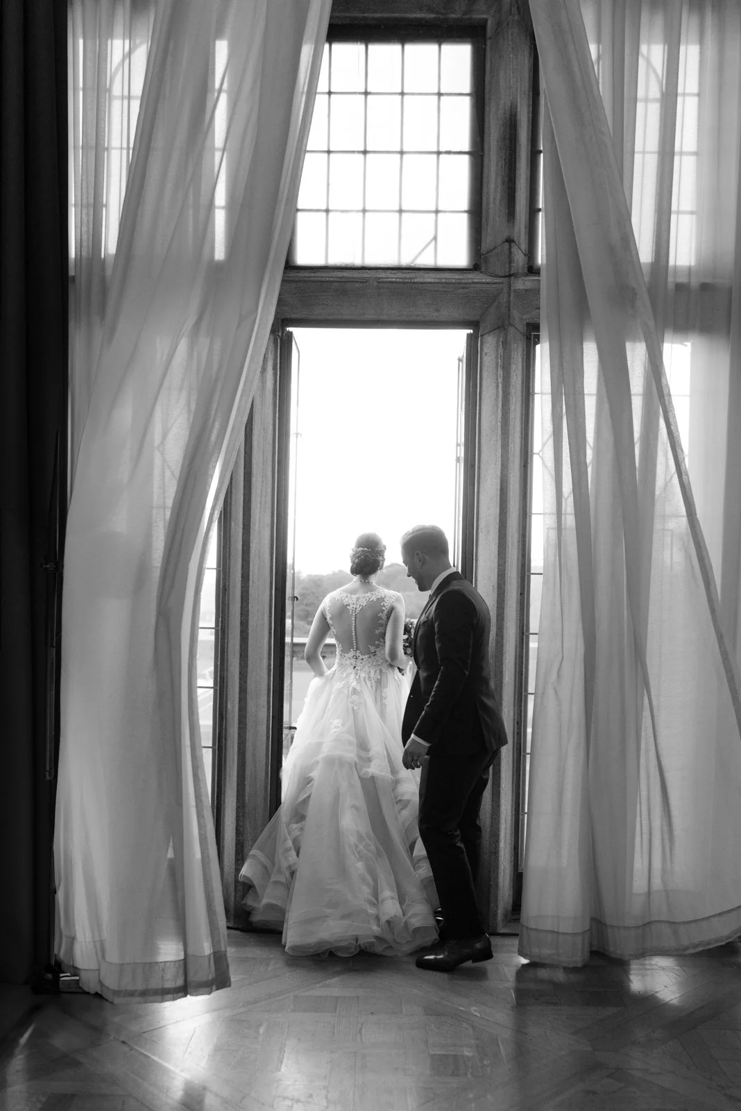 Black and white photo of a bride and groom standing by a large window with sheer curtains. The bride is in a wedding dress facing away, and the groom is in a suit facing the bride, holding hands.