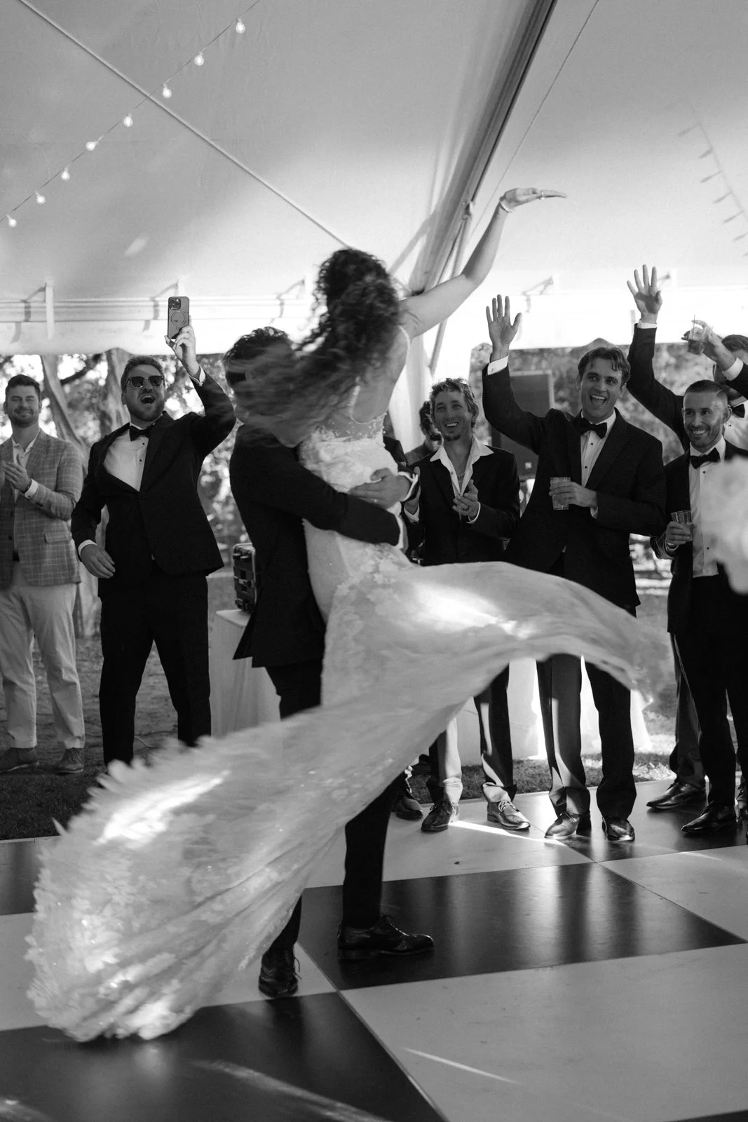Black and white photo of a wedding reception showing a groom lifting the bride while dancing, surrounded by guests happily watching and cheering under a canopy with string lights.
