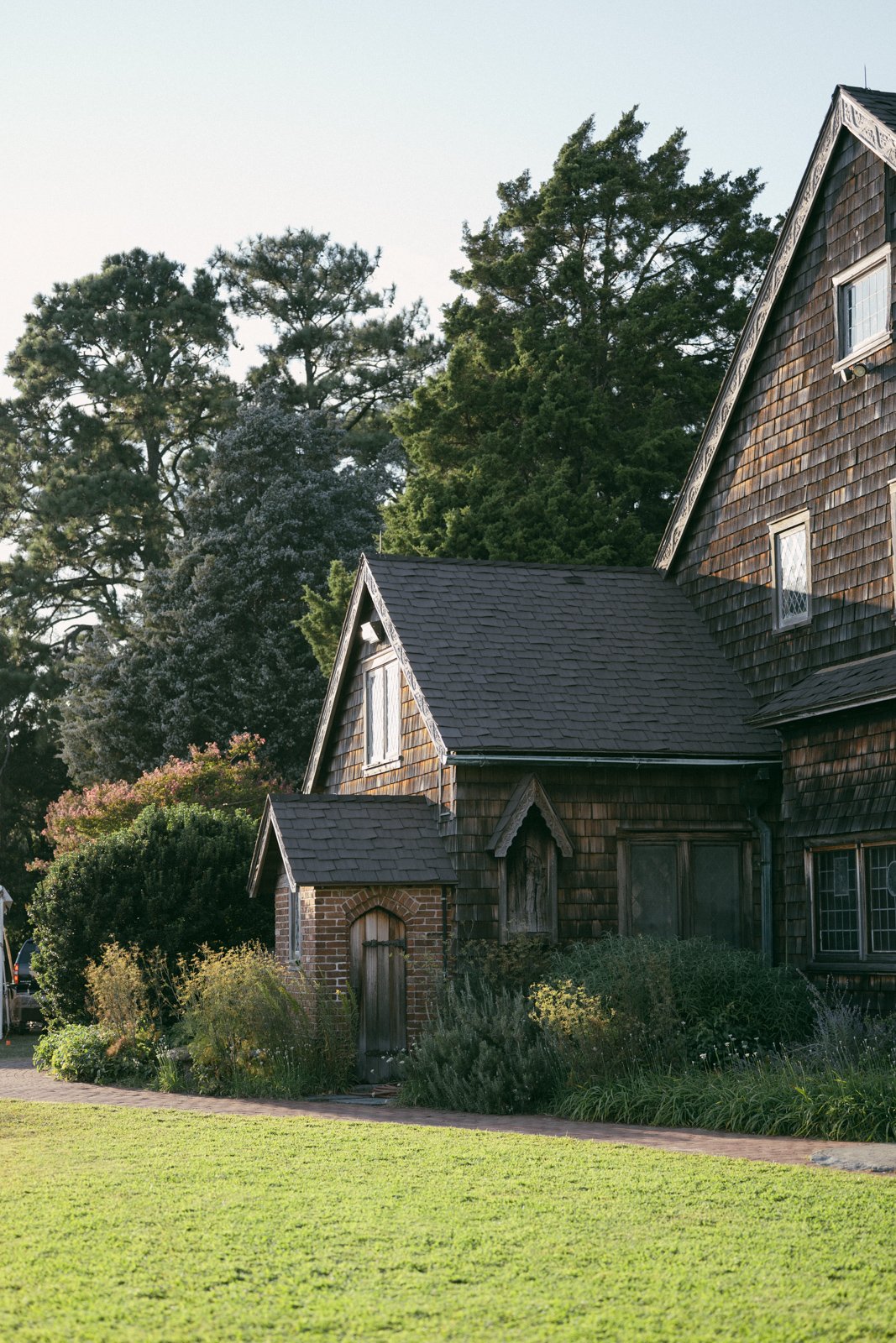 A rustic house with shingle siding and a small brick archway door, surrounded by lush bushes and grass, with tall trees in the background under a clear sky.
