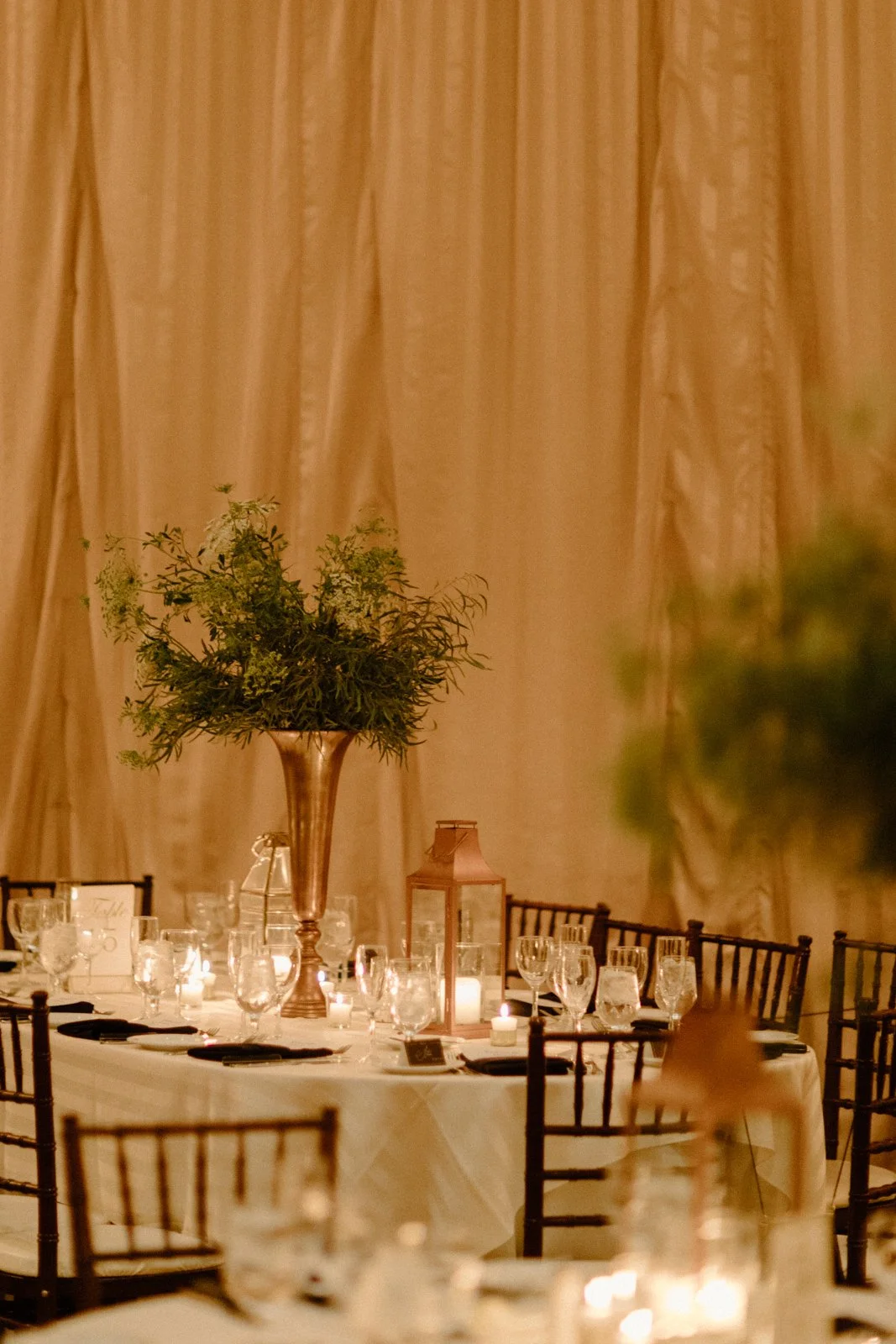 A table set for a formal event, decorated with candles, glasses, and a large floral centerpiece in a tall metallic vase, with beige curtains in the background.