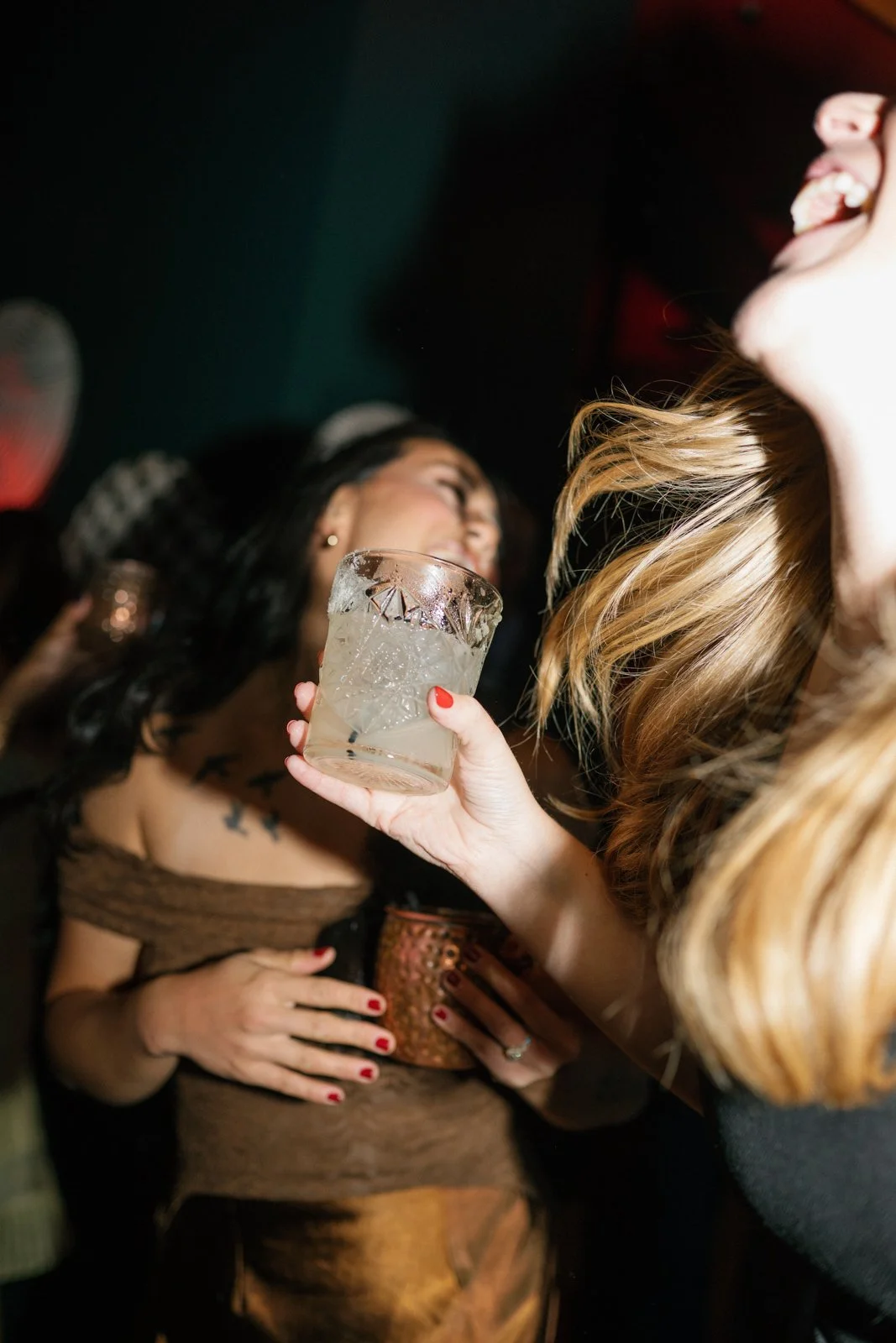 Two women at a party, one woman with blonde hair holding a drink close to another woman with dark hair, revealing her tattooed shoulder, in a dimly lit setting.