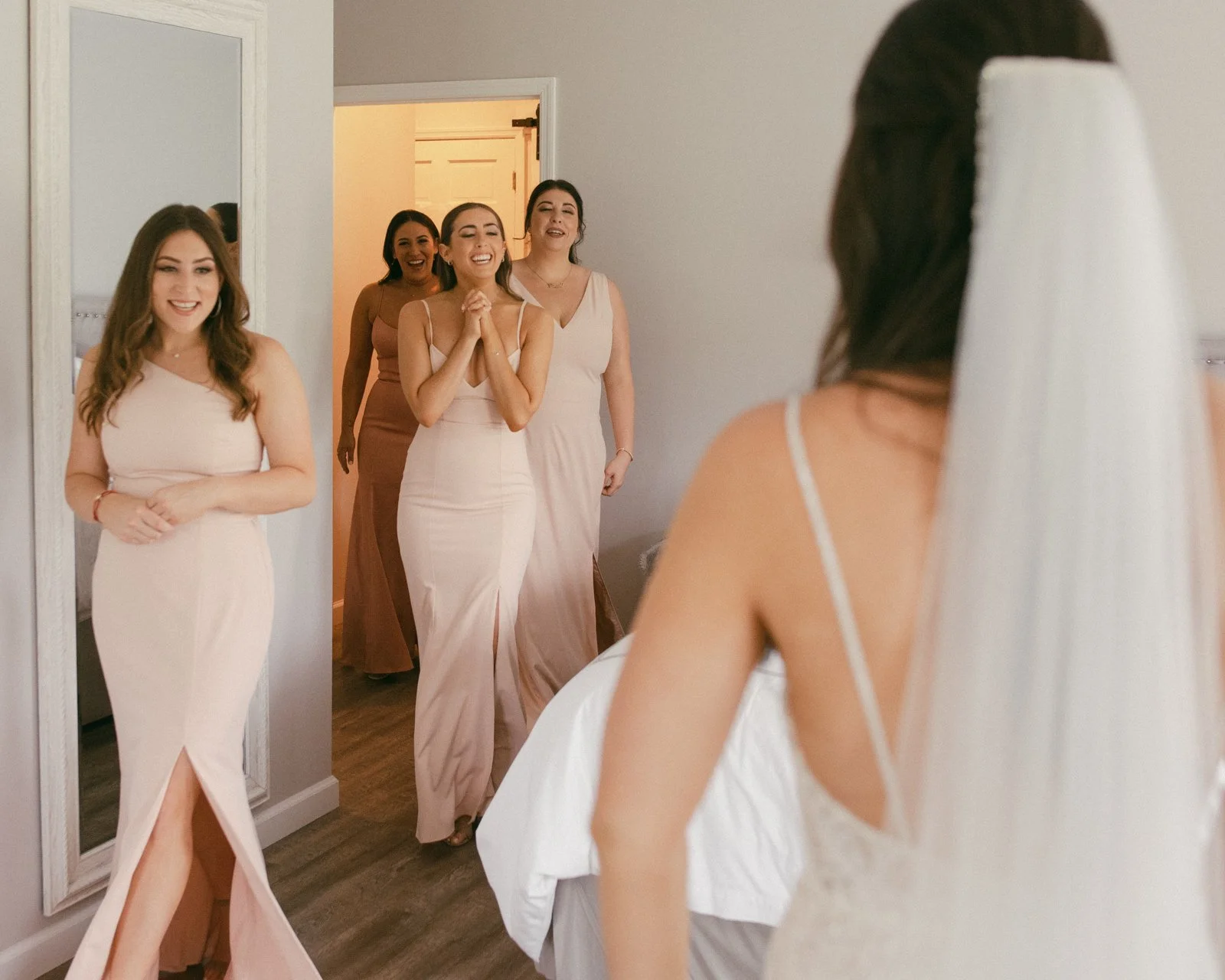 Five women in pastel-colored dresses, one with a veil, are in a room with a mirror, smiling and reacting happily to the bride who is in the foreground.
